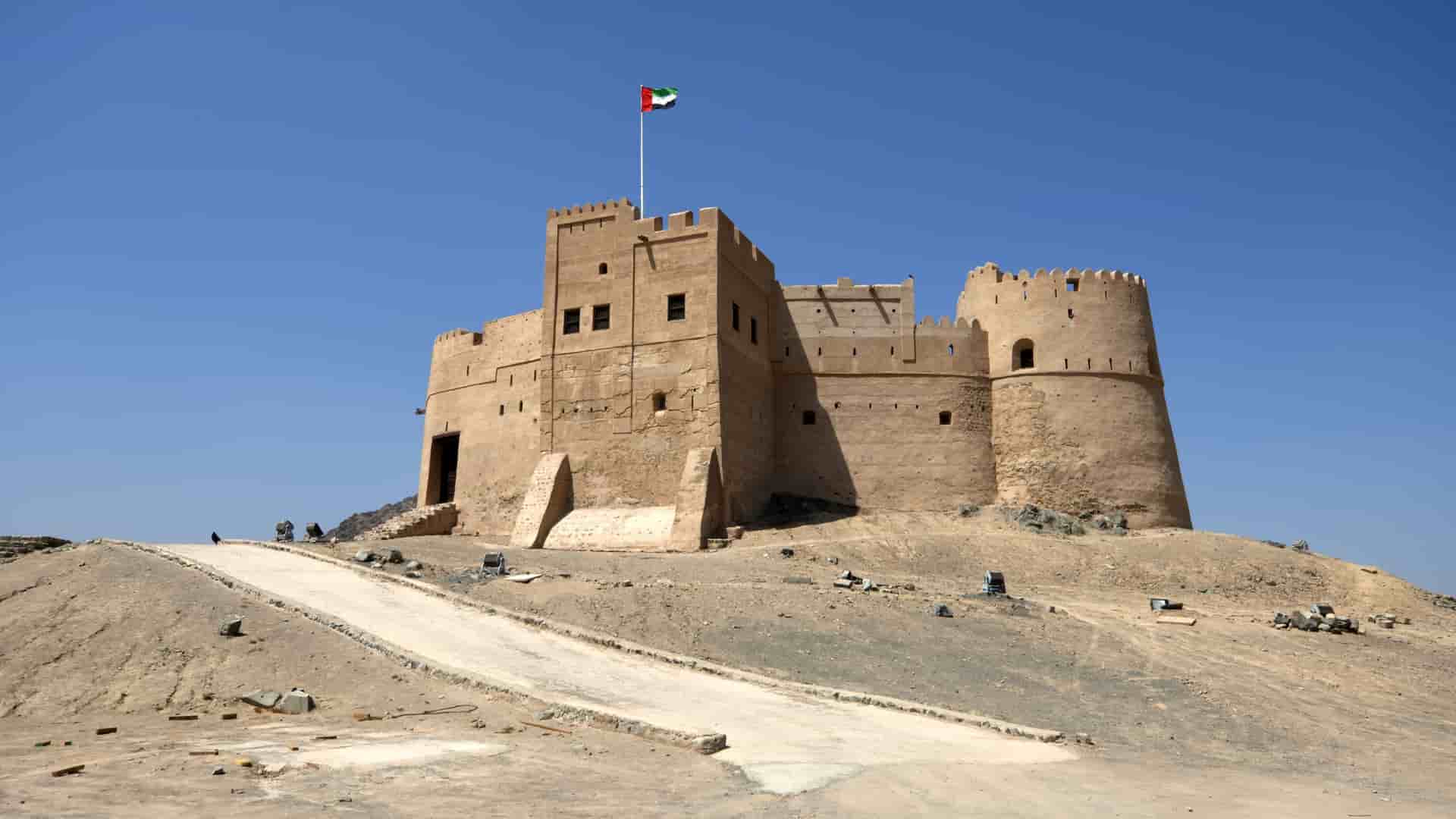 A large, ancient mudbrick fort with a tall central tower and turrets, Fujairah Fort, perched on a rocky hill under a clear blue sky, with a flag of the United Arab Emirates waving on top.