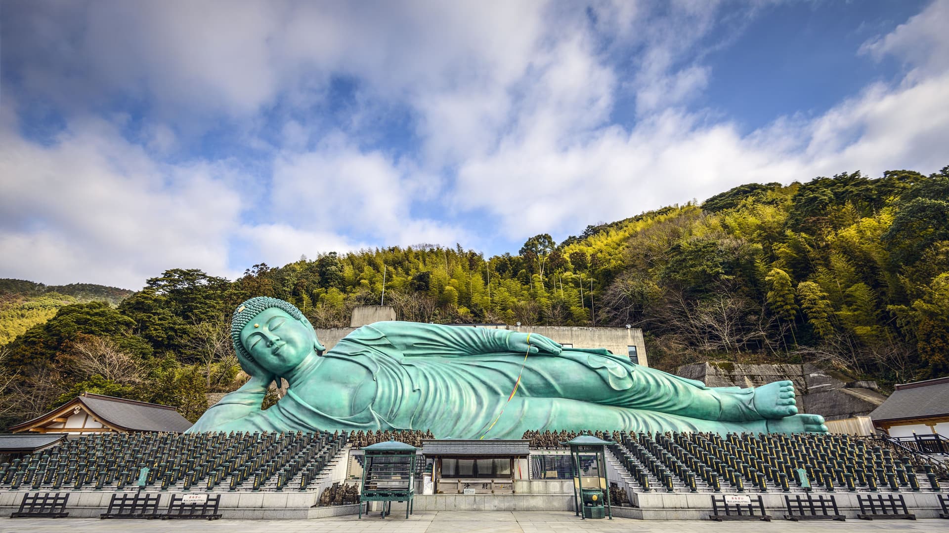A panoramic shot of the enormous, reclining bronze Buddha statue at Nanzoin Temple in Fukuoka, Japan, with green trees and a building in the background.