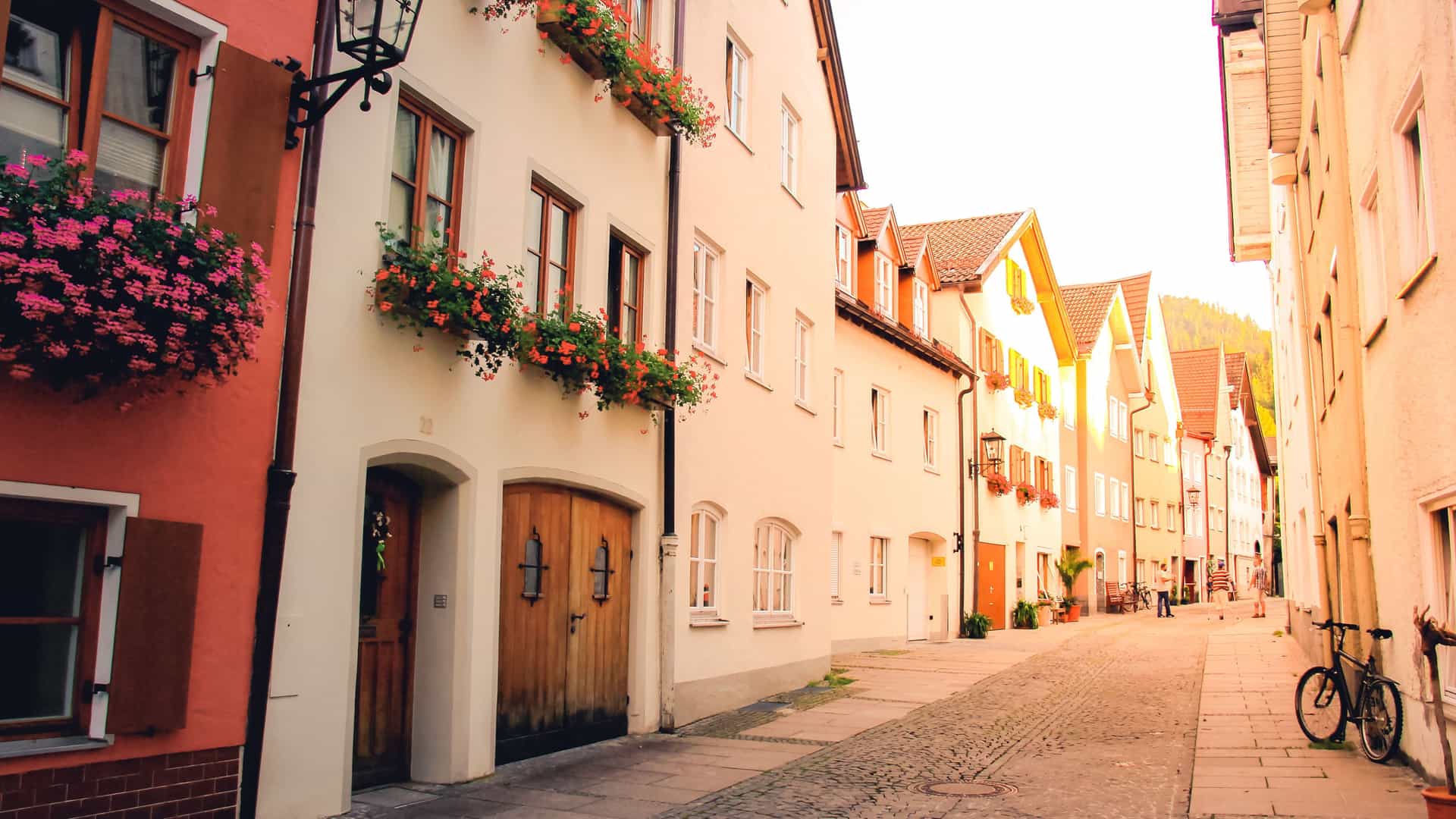 "A narrow, cobbled street in the charming town of Füssen, Germany, with a line of traditional buildings with bright window boxes full of red flowers and a person with a bicycle. "