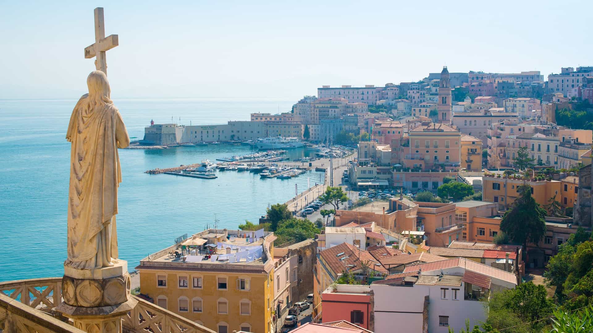 A panoramic view of the coastal city of Gaeta, Italy, taken from the elevated terrace of the Temple of San Francesco. In the foreground, a stone statue, likely St. Francesco or a related figure, stands overlooking the scene. The medieval town, with its dense collection of buildings and monuments, stretches towards the port where ships are docked in the bright blue Tyrrhenian Sea.