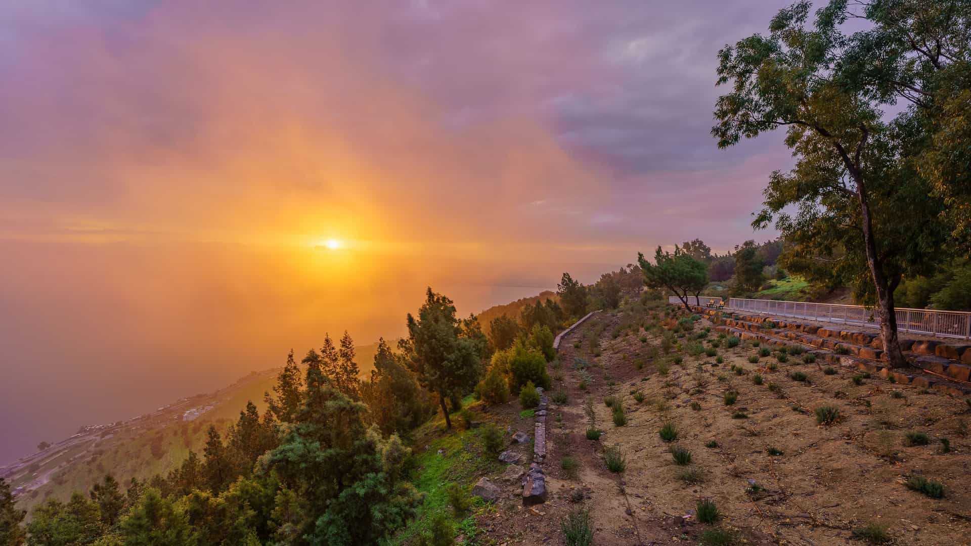 "A dramatic sunset view over the Sea of Galilee in Israel, with the sun casting a golden glow through the clouds as it sets behind the hills with trees and a path in the foreground. "