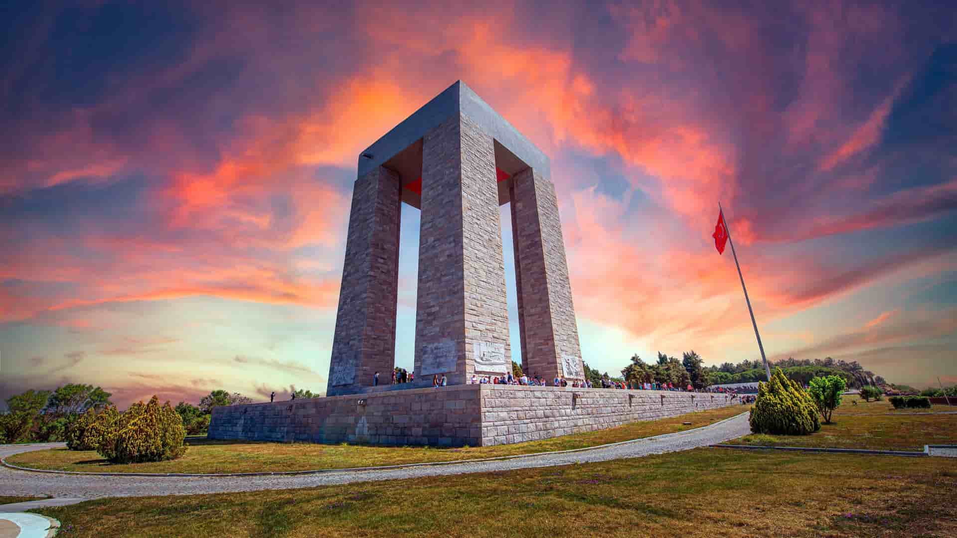 A striking wide-angle shot of the Canakkale Martyrs' Memorial in Gallipoli, Turkey, a massive stone monument honoring fallen soldiers, silhouetted against a dramatic and colorful sunset sky.