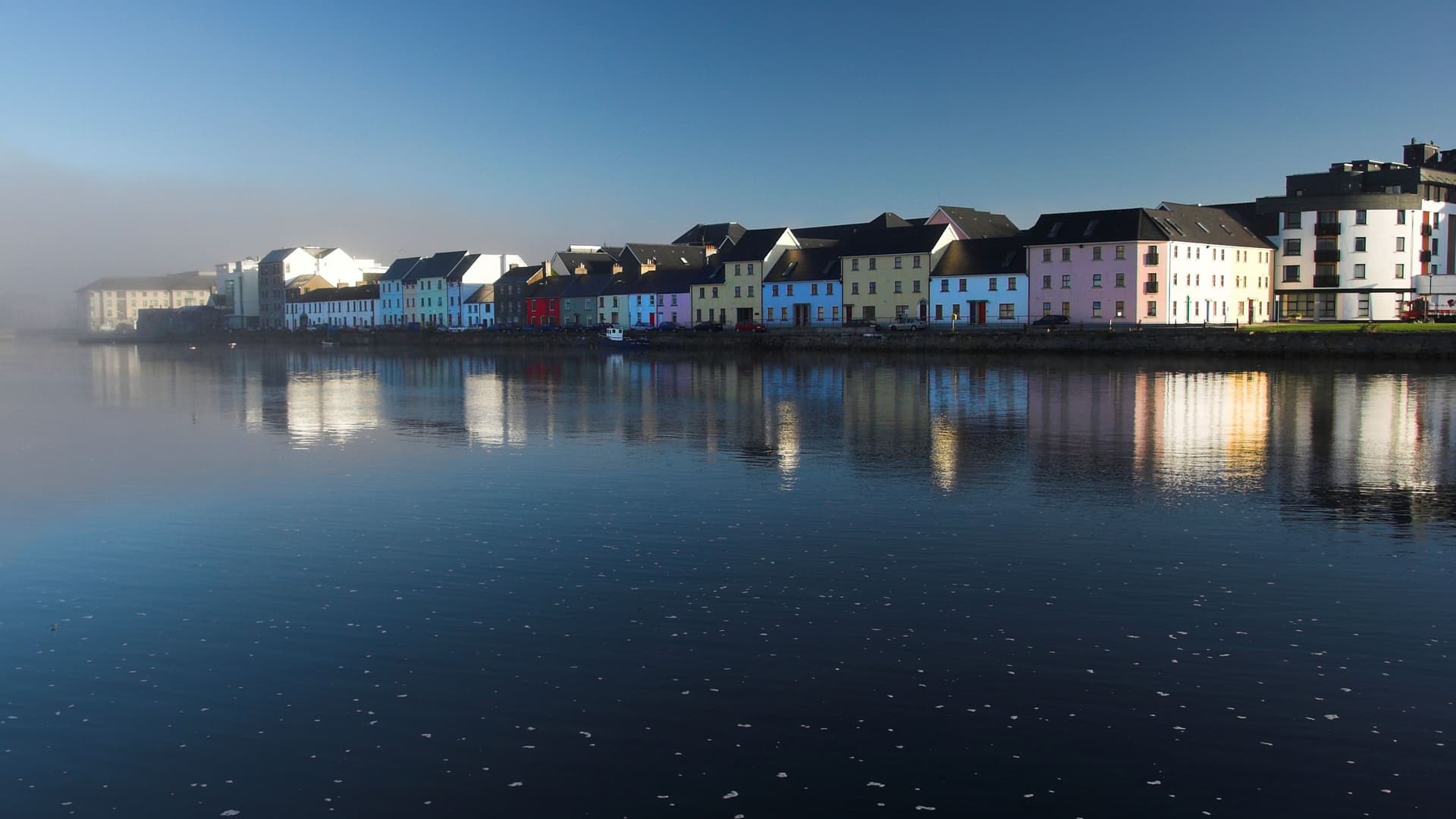 A tranquil shot of a row of colorful houses lining the waterfront in Galway, Ireland, their reflections shimmering on the calm surface of the Corrib River on a foggy day.
