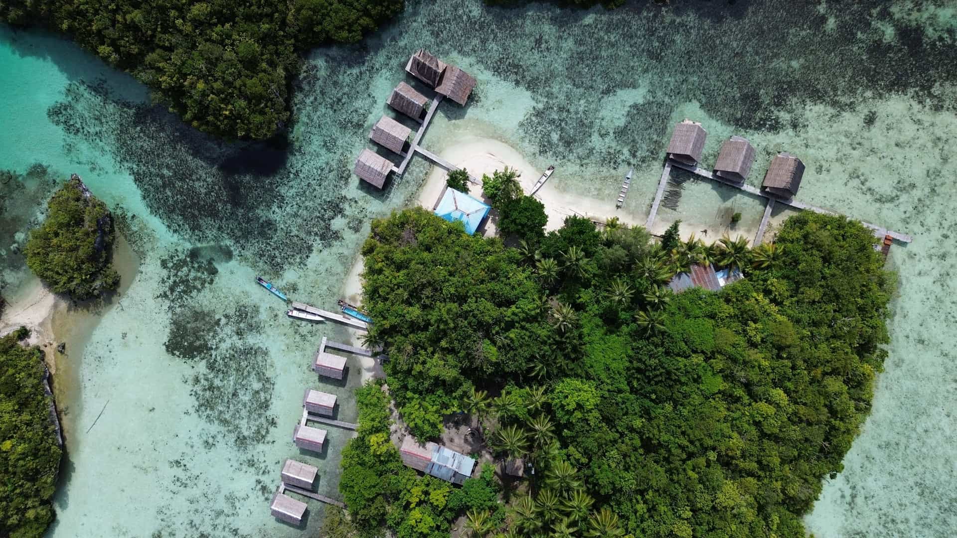 An aerial shot of Gam Island, Indonesia, showing a cluster of rustic bungalows built on stilts over the crystal-clear turquoise water, surrounded by lush green tropical foliage.