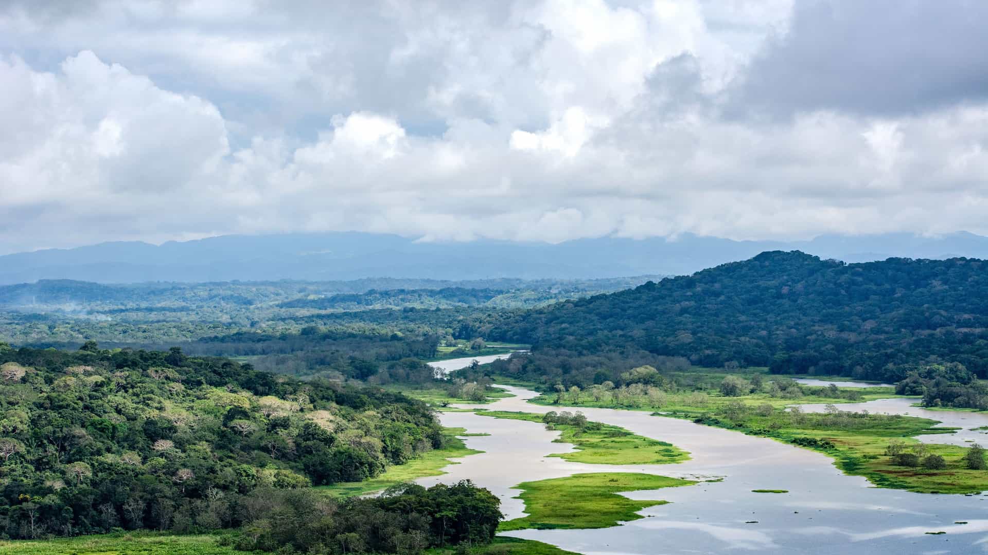 "A scenic aerial shot of the Chagres River winding through the dense, green rainforest of Gamboa, Panama, with mountains in the misty distance and a cloudy sky above. "