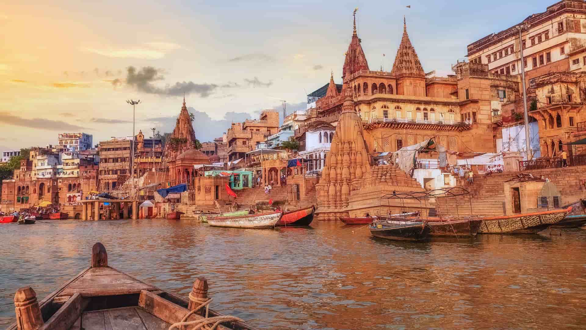 "A wide-angle view of the Ganges River in Varanasi, India, with a row of ancient buildings, temples, and ghats descending to the water's edge, all under a warm evening sky. "