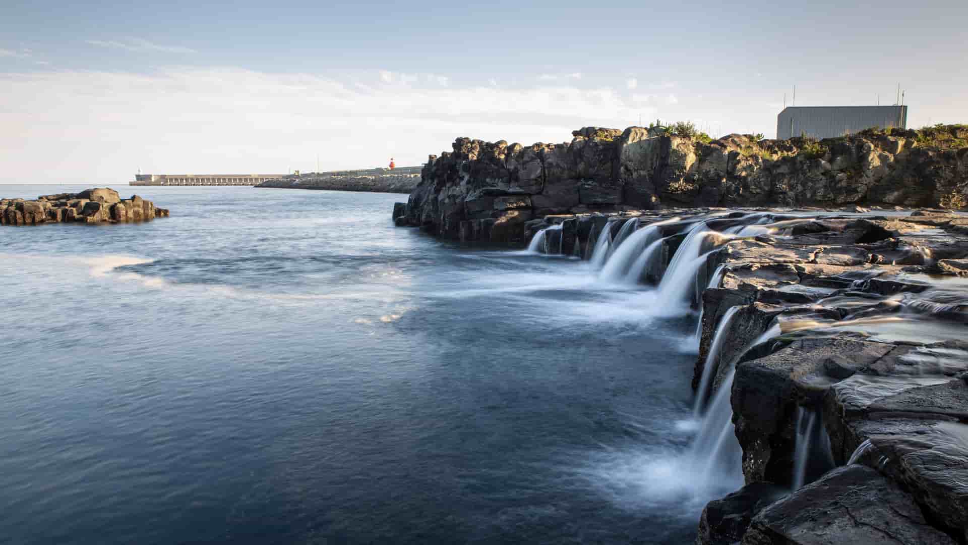 A serene long exposure shot of a waterfall cascading over dark volcanic rock into the calm sea in Gangjeong, South Korea, with a breakwater and lighthouse visible in the distance.
