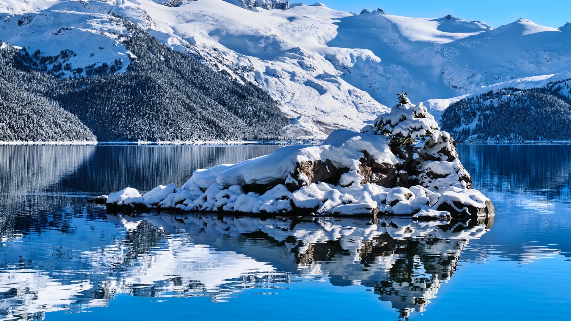 A beautiful mountain lake with a small, snow-covered island in the foreground, with majestic, snow-capped mountains and evergreen trees in the background under a clear blue sky.