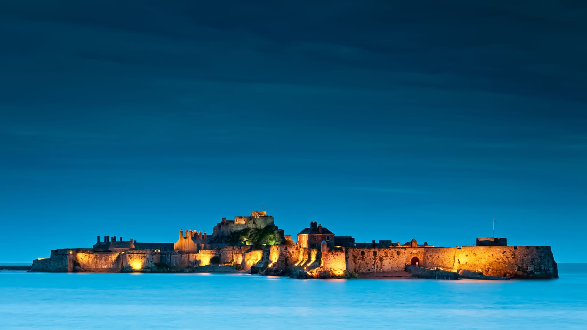 A historic castle fortress illuminated at night, located on a small island surrounded by calm water under a deep blue evening sky.