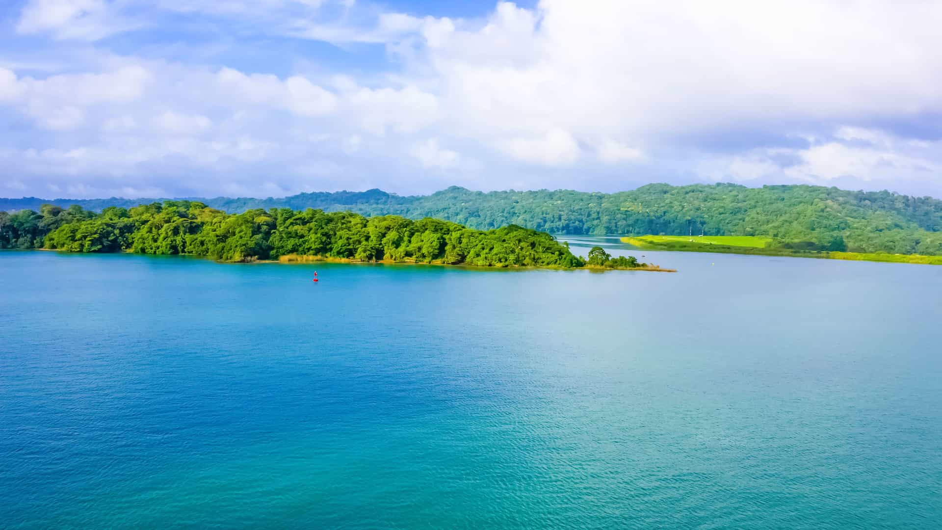 A serene aerial shot of Gatun Lake in Panama, featuring a large body of water surrounded by lush, green, forested islands and distant hills under a partly cloudy blue sky.