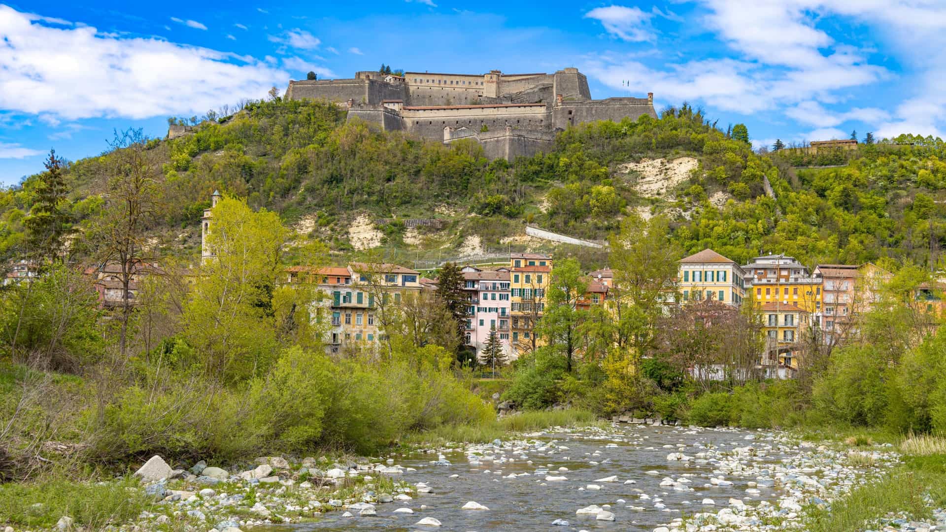 The medieval Forte di Gavi (Gavi Fortress) positioned on a hilltop, overlooking the colorful buildings of the town of Gavi, Piedmont, Italy, with a rocky riverbed and stream in the foreground.