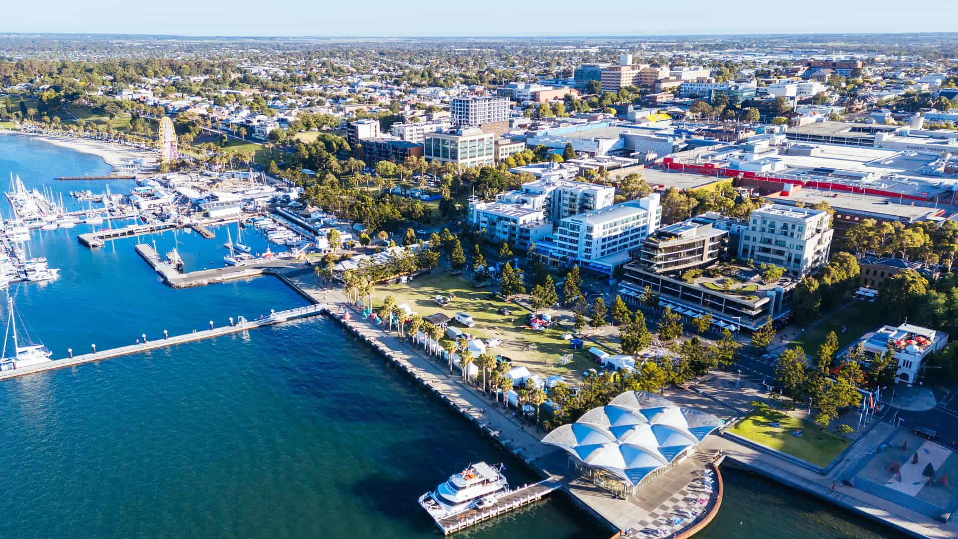An aerial view of the vibrant waterfront of Geelong, Australia, with a marina full of boats, a bustling city skyline, and a Ferris wheel in the background.