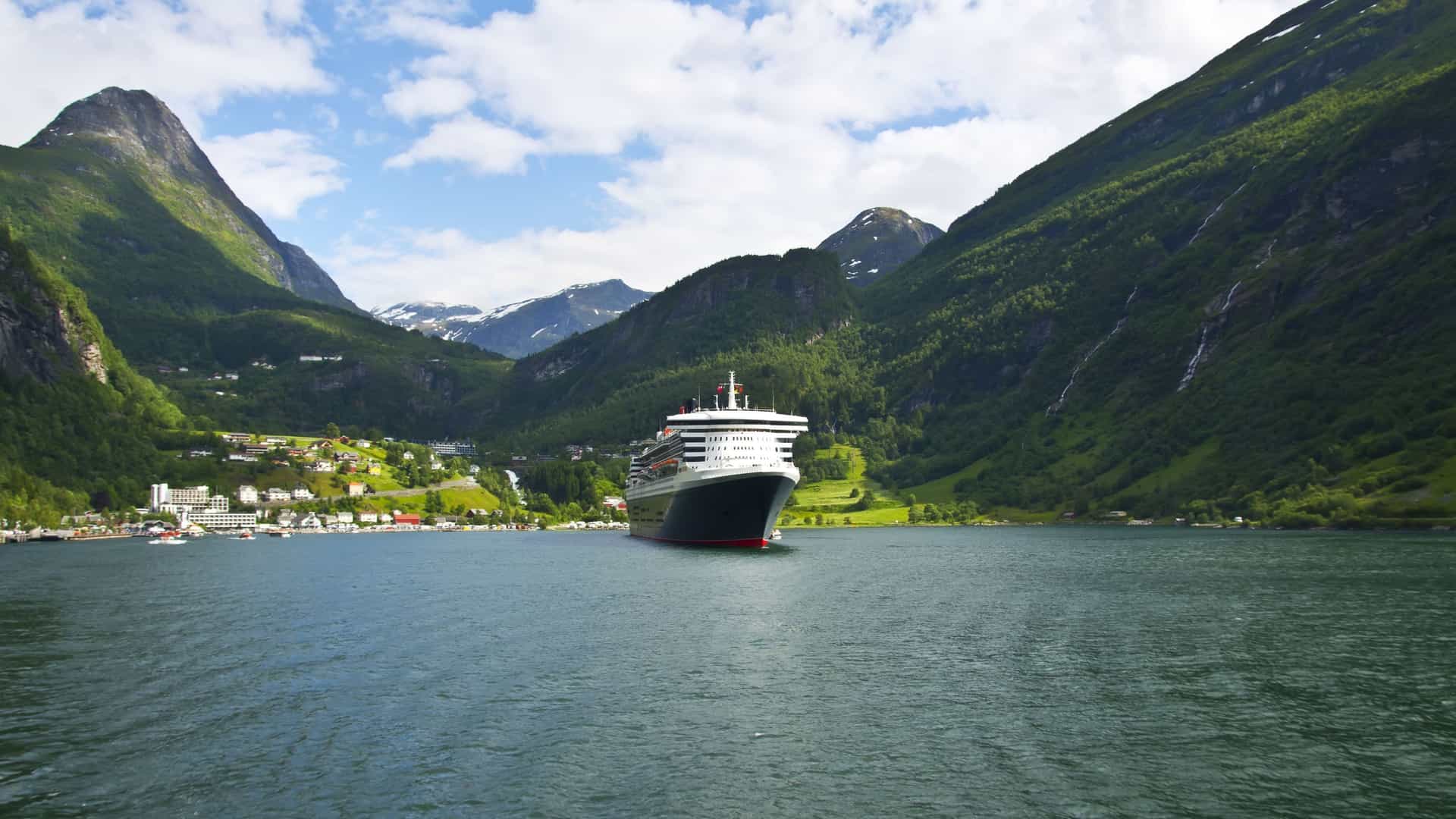 A stunning view of a large cruise ship anchored in the scenic Geirangerfjord, Norway, with towering green mountains and a small village in the background.