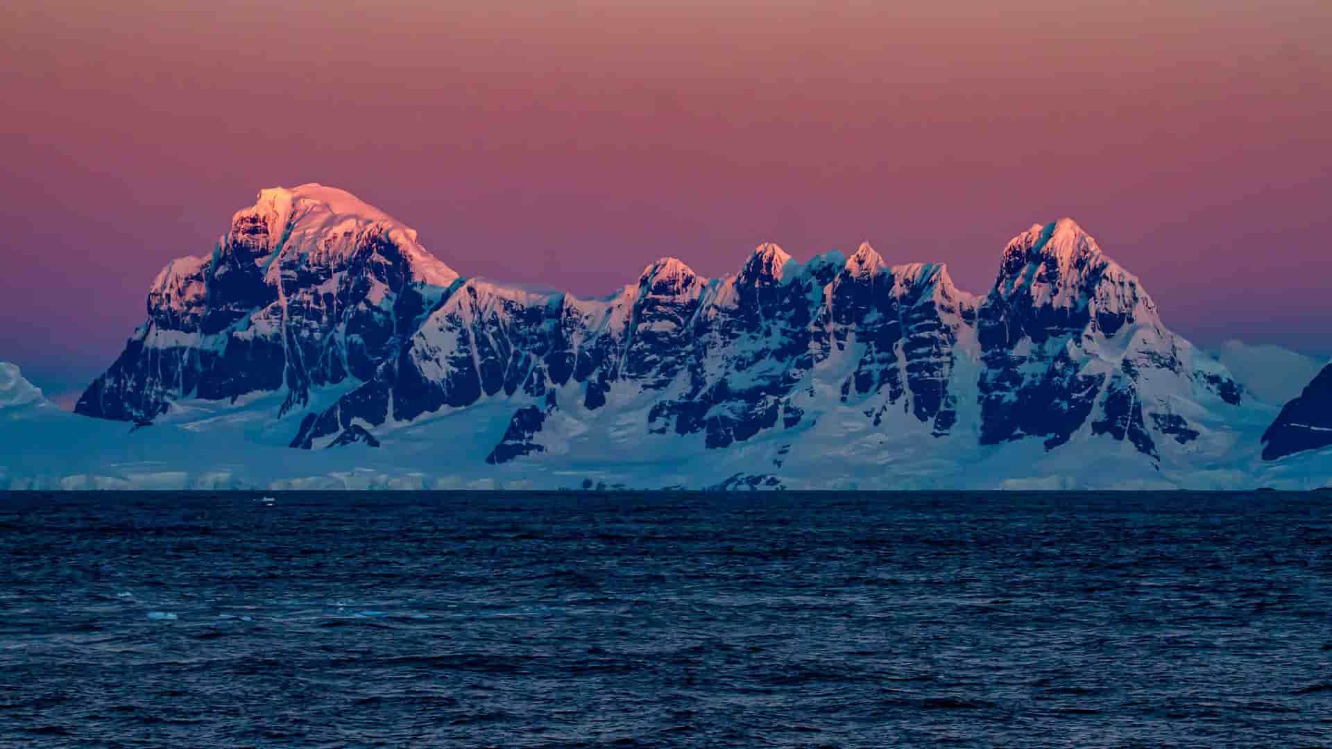 A dramatic sunset view over the Gerlache Strait in Antarctica, featuring snow-covered mountains with vibrant pink and purple hues reflected on the peaks.