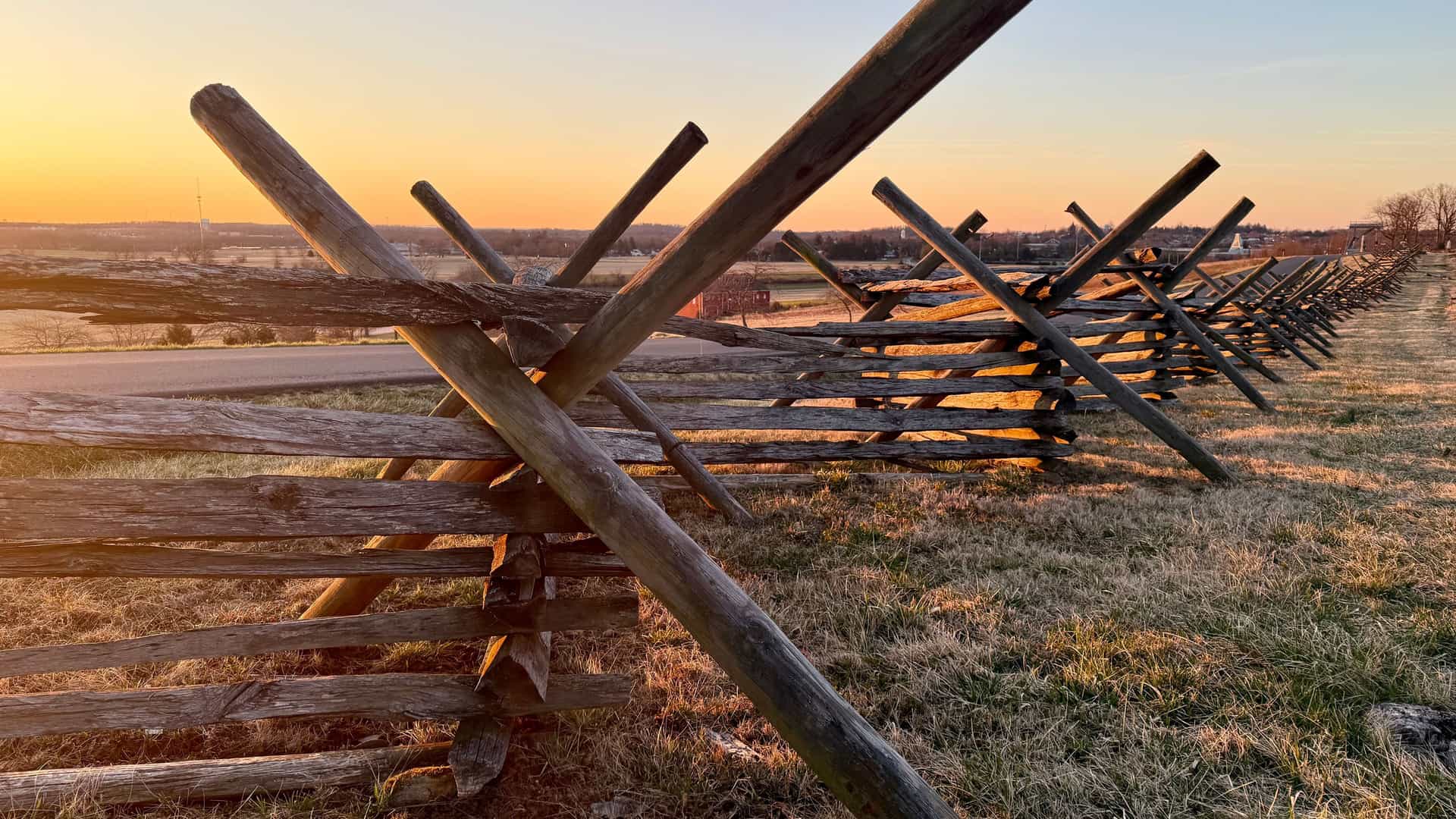 A scenic ground-level shot of a historic wooden split-rail fence on a grassy field at the Gettysburg National Military Park at sunset, with golden light illuminating the landscape.