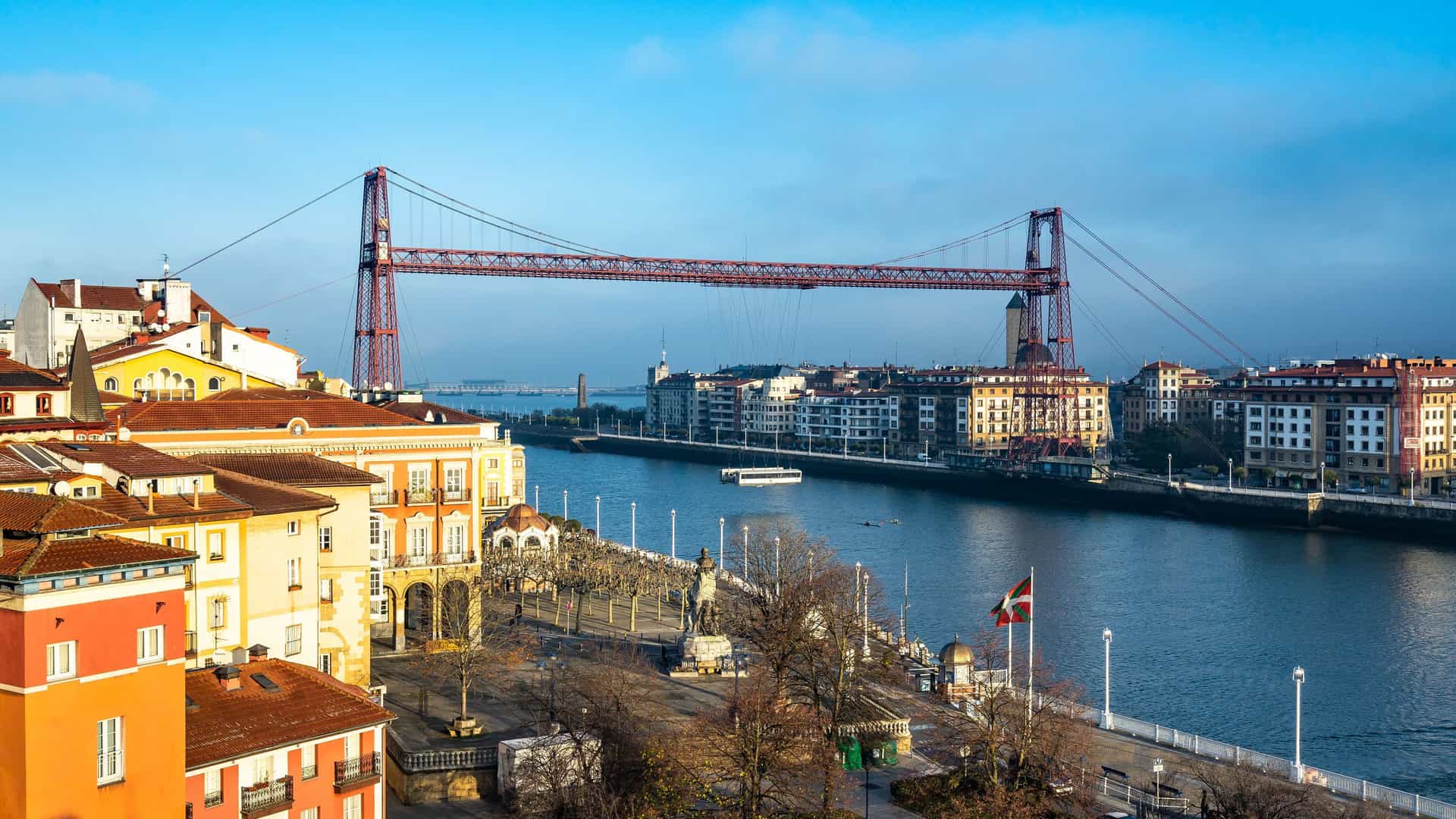 A panoramic shot of Getxo, Spain, featuring the UNESCO-listed Vizcaya Bridge over the Nervión River with historic buildings and a statue in the foreground.