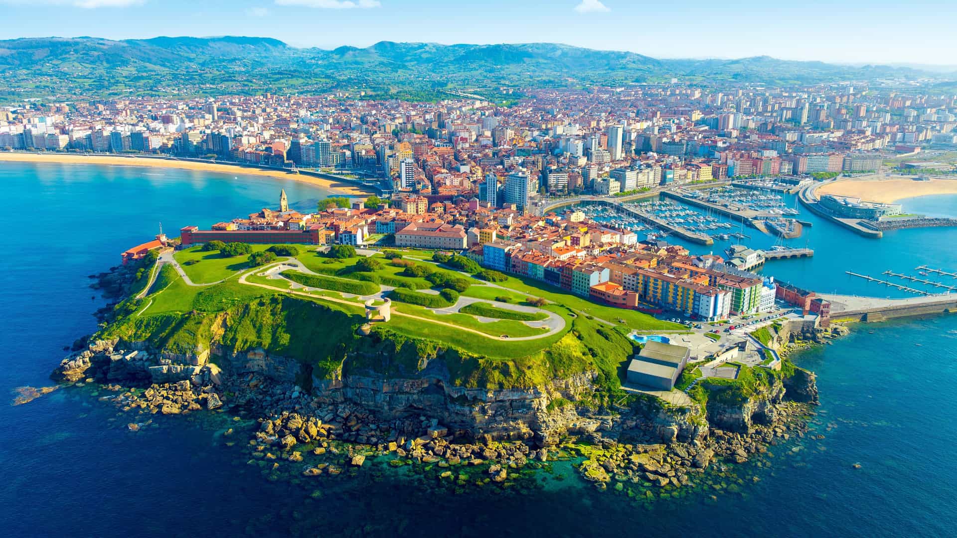 An aerial shot of Gijon, Spain, showcasing the Cimavilla Peninsula with green grass and cliffs, and the beautiful city and marina in the background.