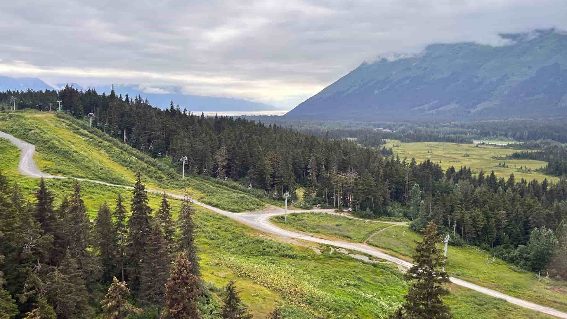 "A high-angle view of the scenic landscape of Girdwood, Alaska, with ski slopes covered in green grass in the foreground, and a dense forest leading to a wide river valley with mountains shrouded in mist. "