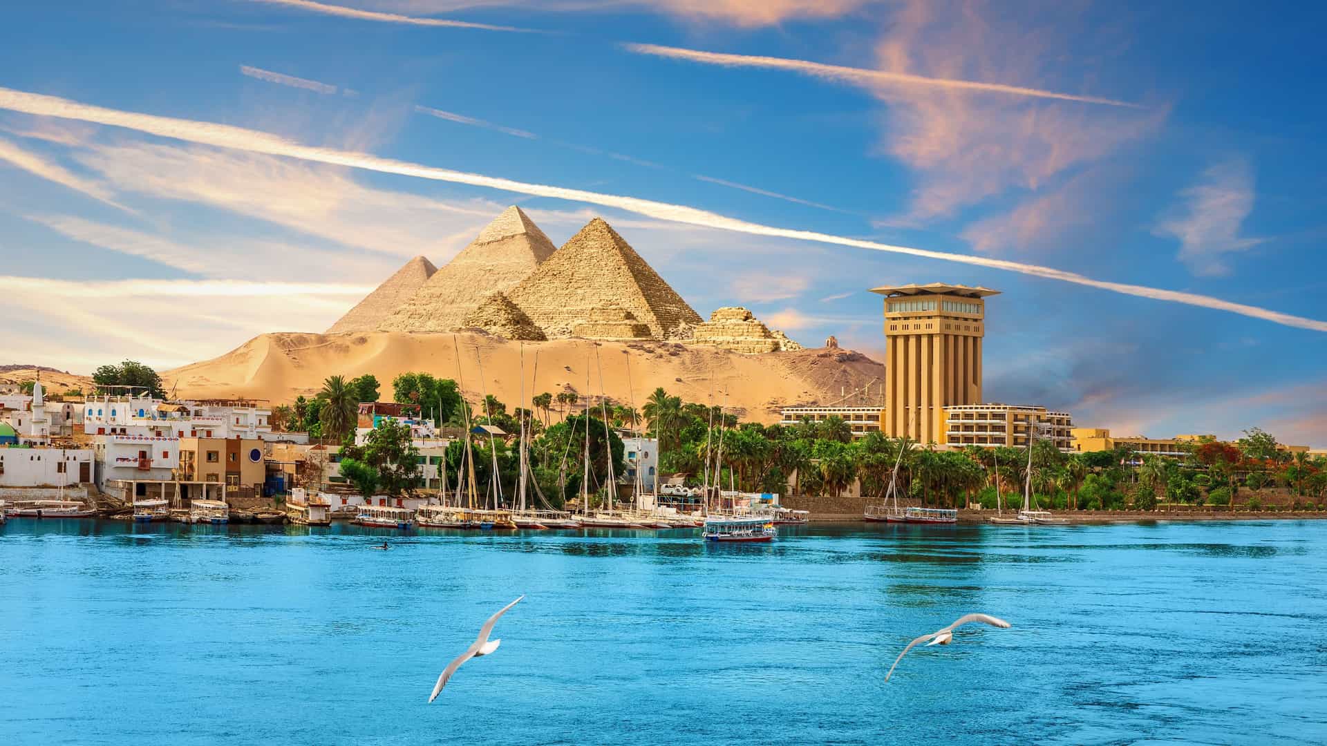 A wide-angle landscape shot of the iconic Pyramids of Giza, Egypt, overlooking the Nile River, with sailboats and buildings in the foreground and seagulls flying above the water.