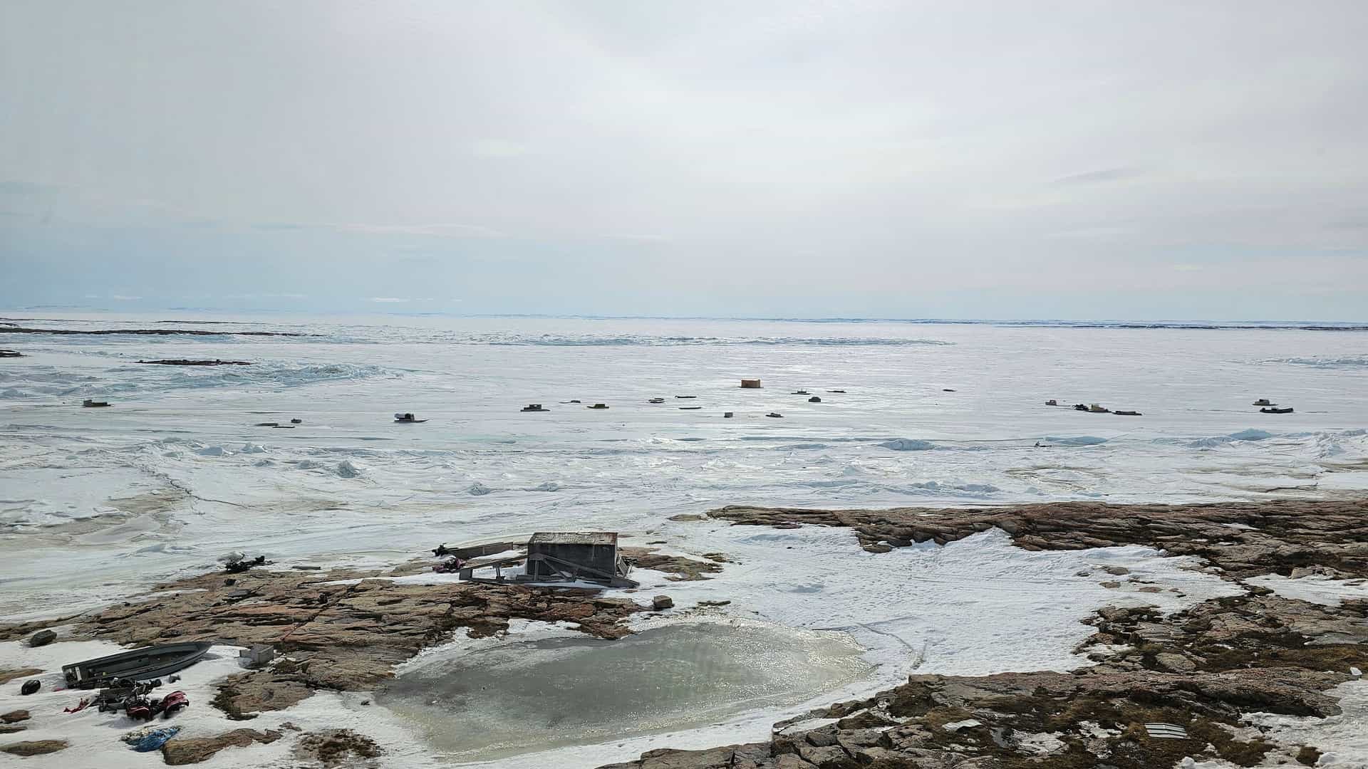 A high-angle view of a frozen bay in Gjoa Haven, Nunavut, showing small ice fishing huts scattered across the vast expanse of ice under a white, cloudy sky.