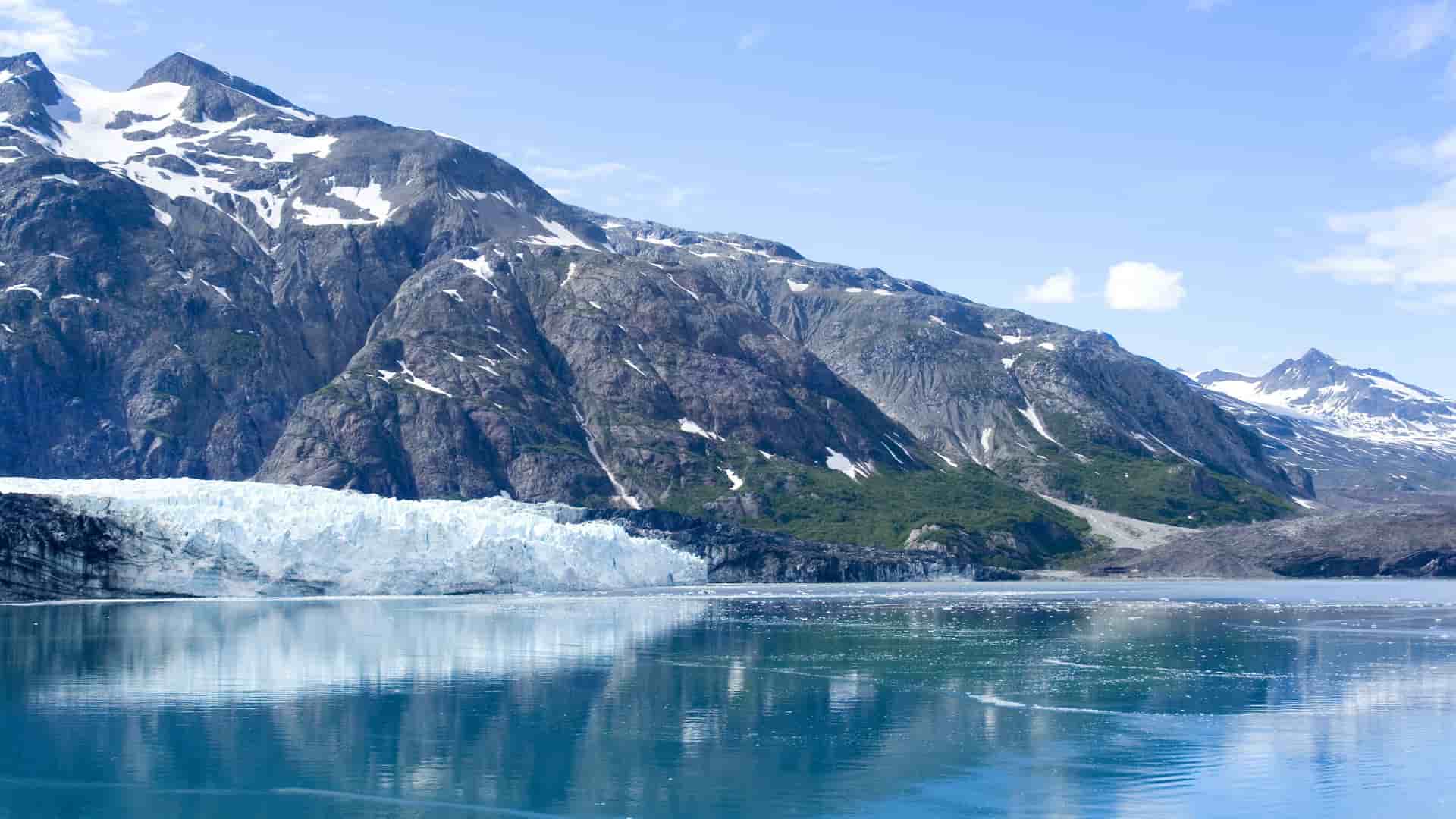 A beautiful landscape of Glacier Bay, Alaska, showing a massive wall of ice and a calm body of water with majestic, snow-capped mountains in the background.