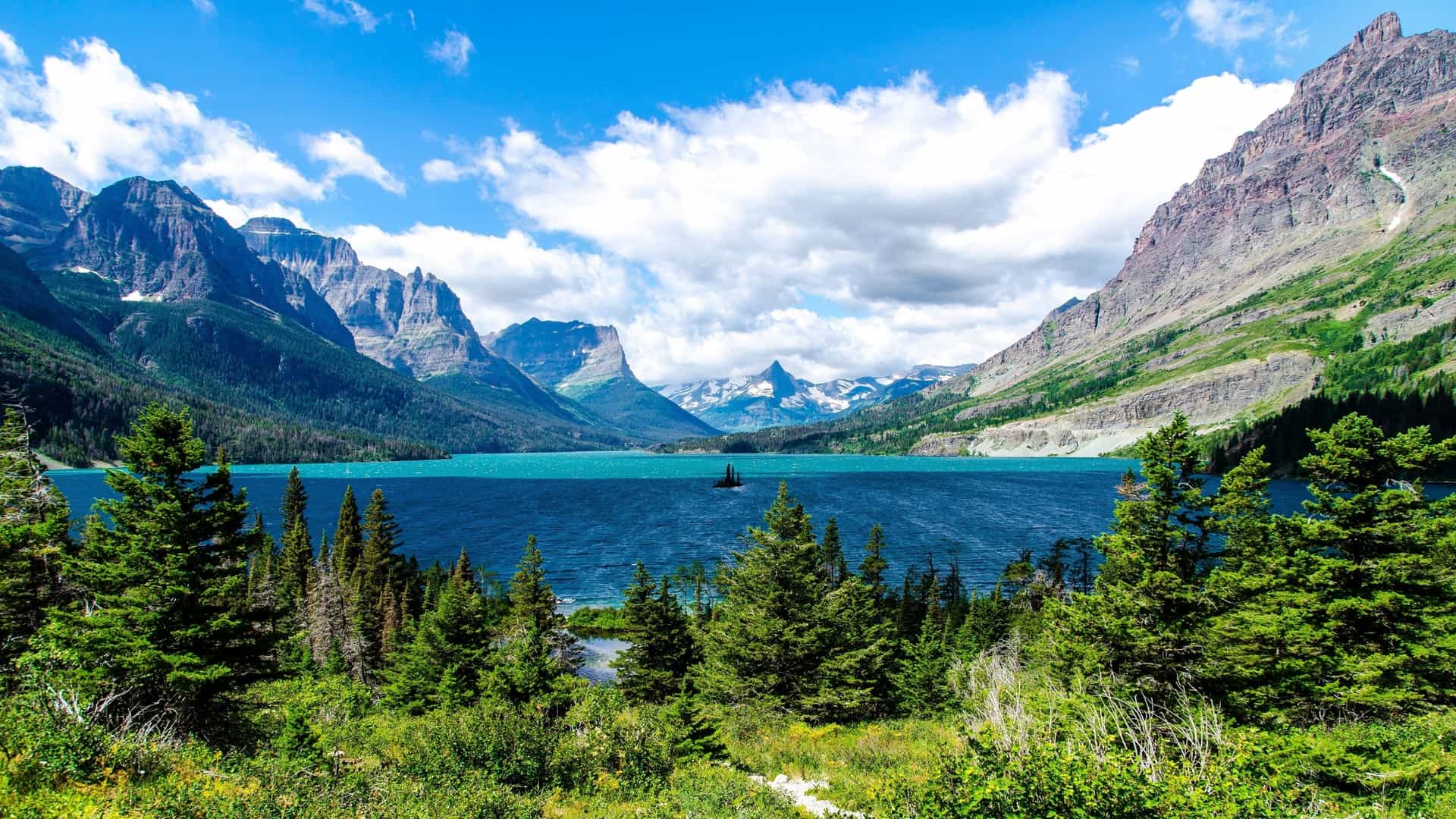 Brightly colored turquoise water of Saint Mary Lake in Glacier National Park, Montana, USA, with steep, rugged mountains and dense evergreen forests under a bright blue sky with clouds.