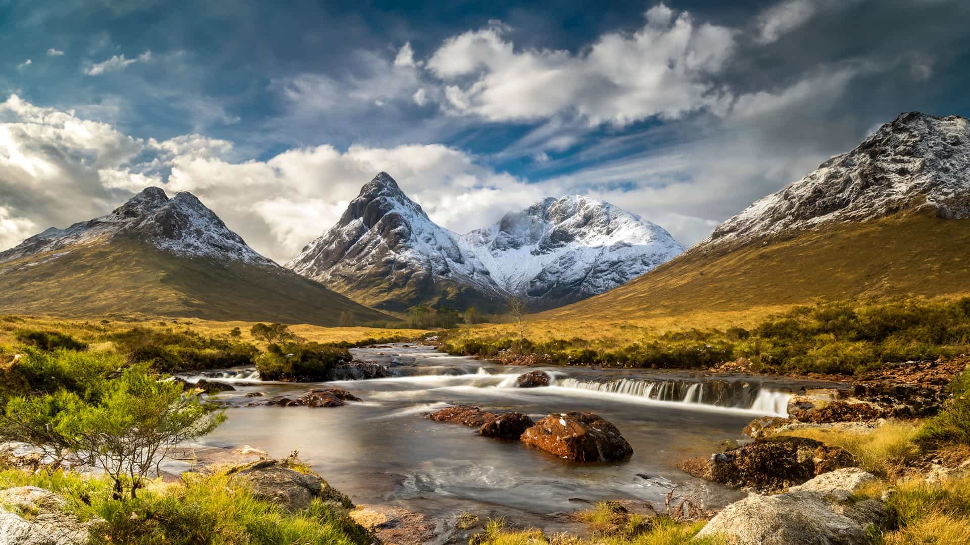 "A dramatic landscape shot of the Glencoe Valley in the Scottish Highlands, featuring snow-capped mountains and a stream rushing over rocks in the foreground under a dynamic, cloudy sky. "