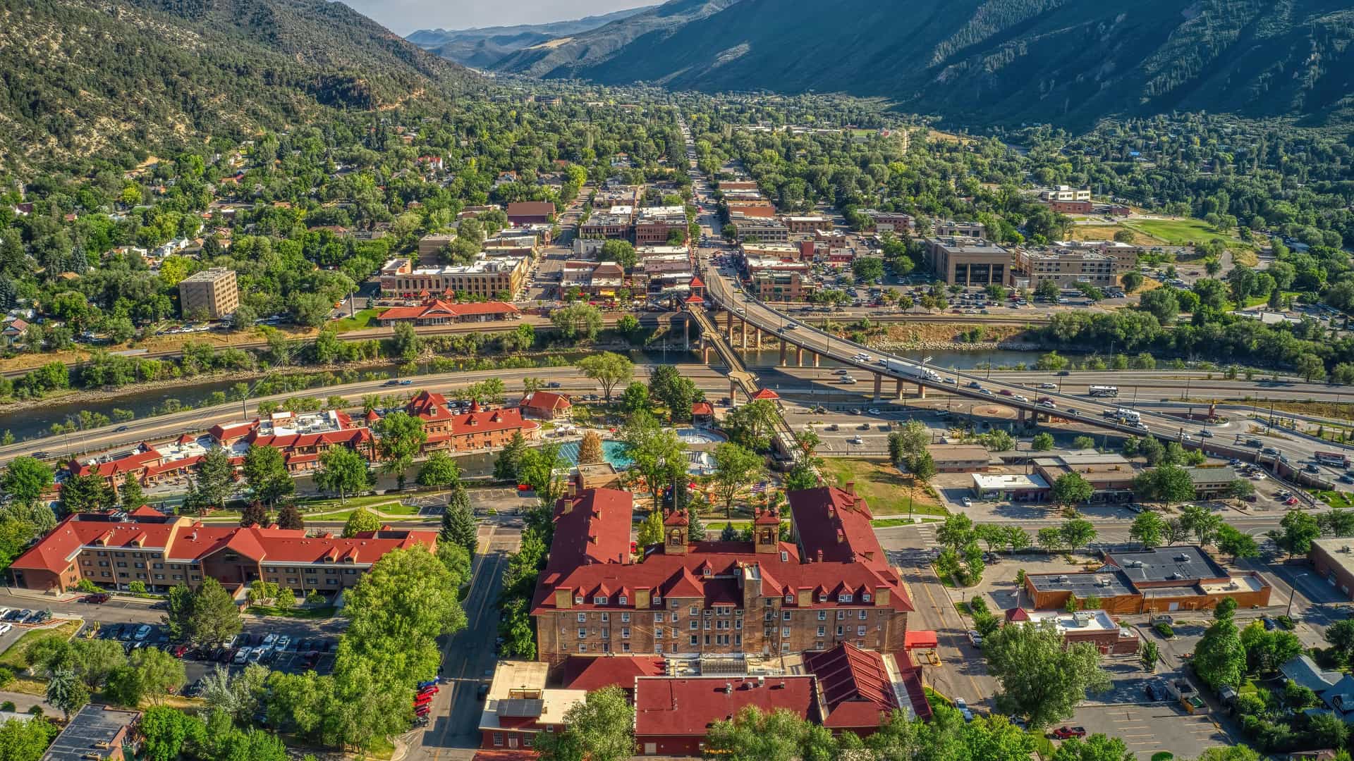 "An aerial view of Glenwood Springs, Colorado, nestled between two mountains, with the prominent Hotel Colorado in the foreground, a highway bridge, and a river flowing through the town. "