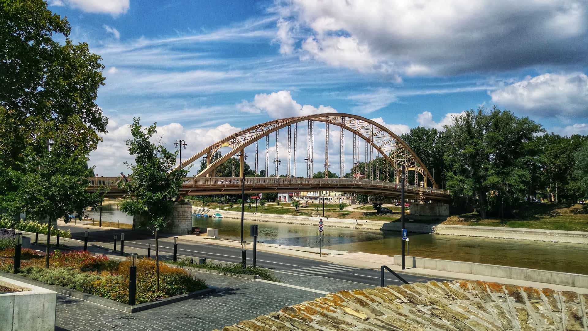 "A wide-angle view of a golden arch bridge crossing a river in Győr, Hungary, with a paved road, trees, and a walkway in the foreground under a bright blue sky. "
