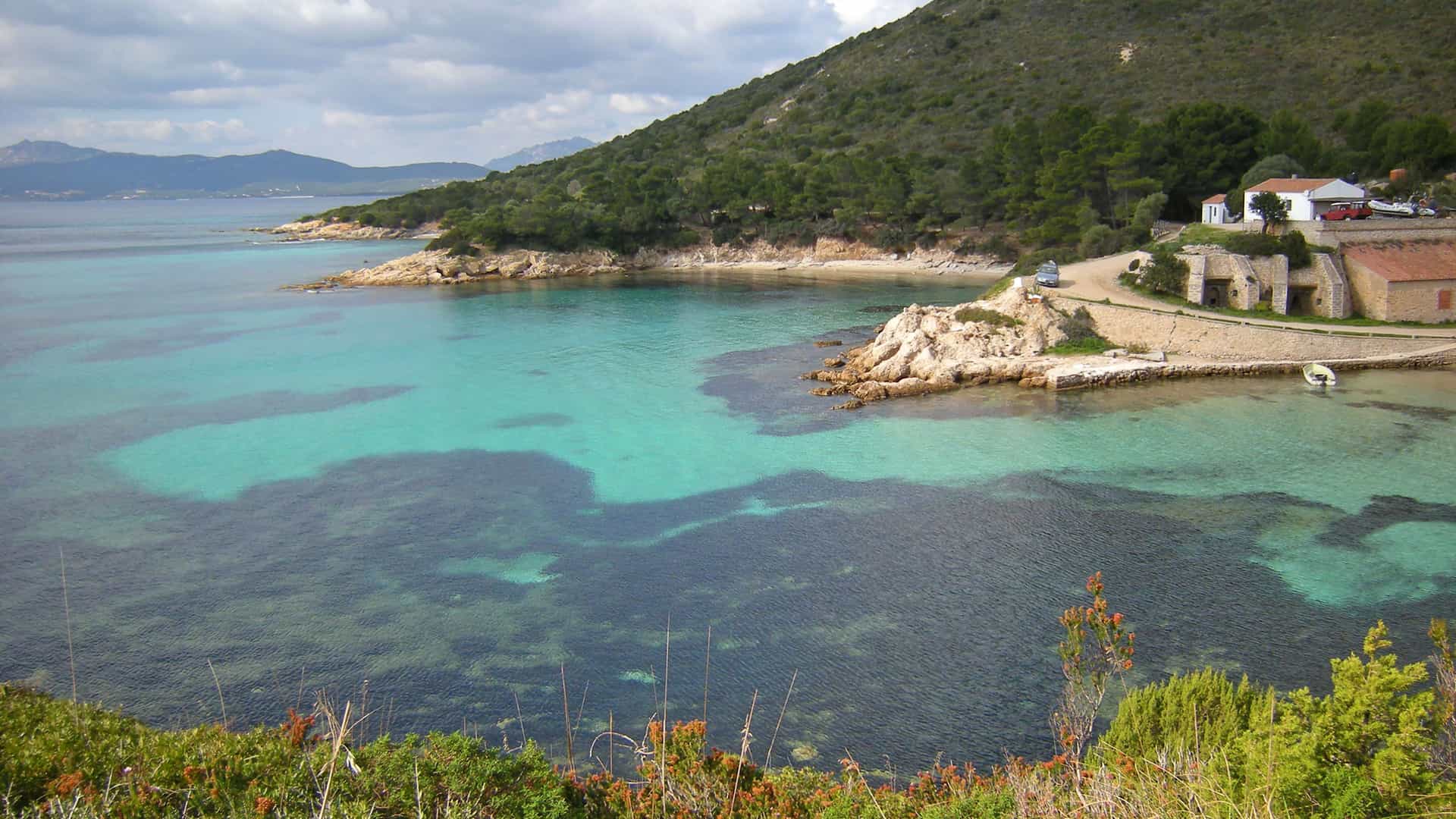 A high-angle view of a serene beach in Golfo Aranci, Sardinia, showcasing the beautiful turquoise water, a sandy shoreline, and a few historic buildings nestled into the green hills.