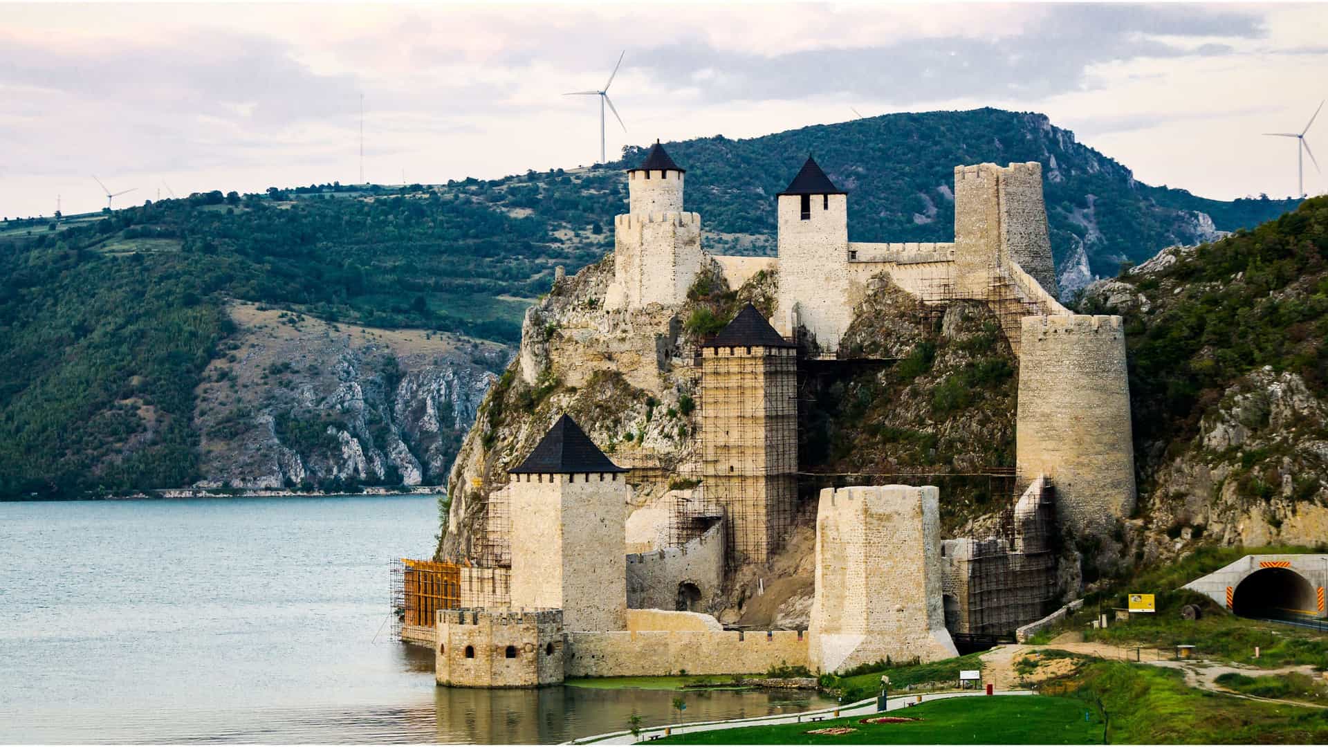 A scenic view of the Golubac Fortress, a medieval castle built on a rocky cliffside by the Danube River, with green mountains and wind turbines in the background.