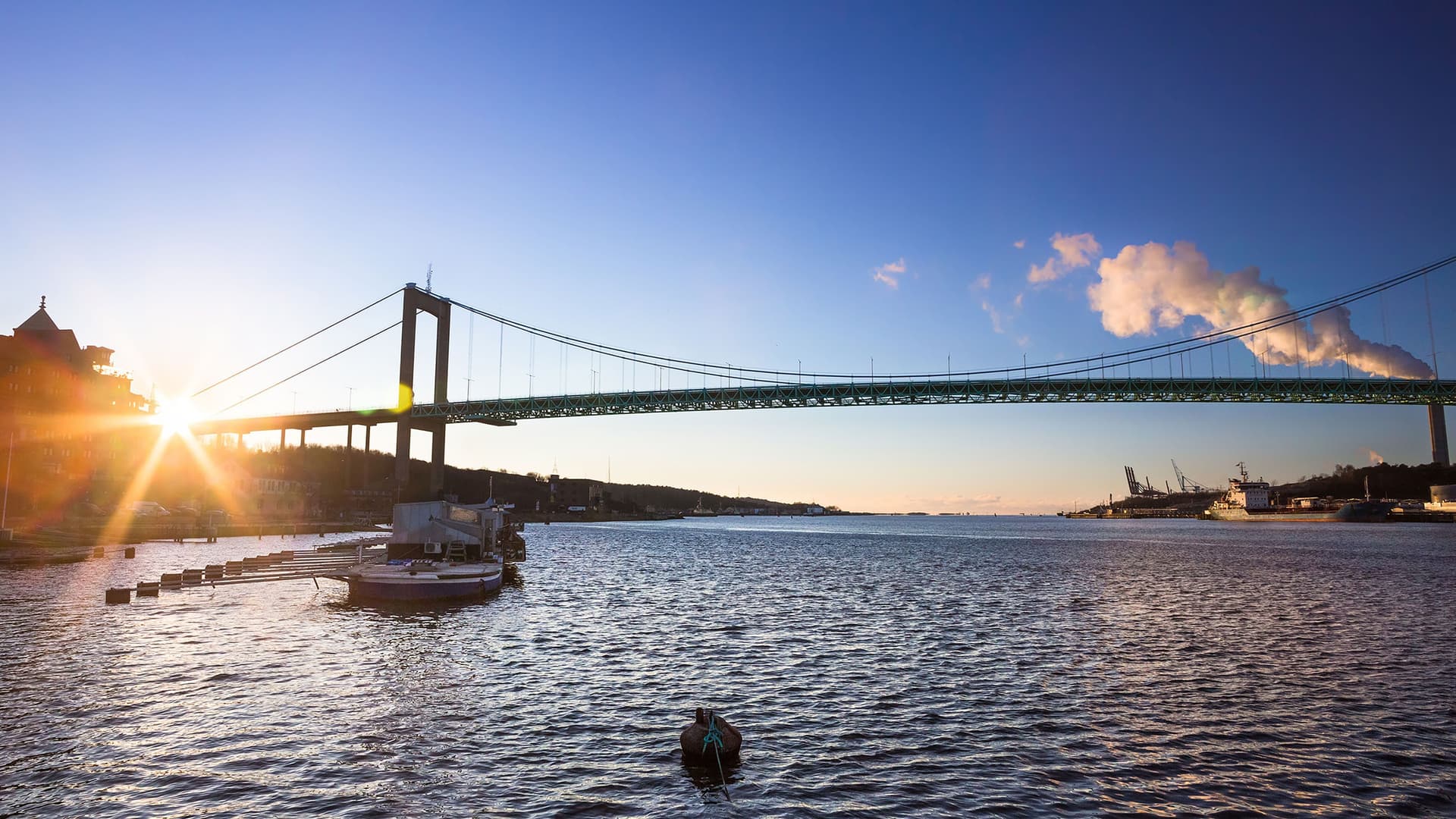 A wide-angle sunset view of the Älvsborg Bridge in Gothenburg, Sweden, spanning over the Göta Älv river with ships and a port visible in the background.