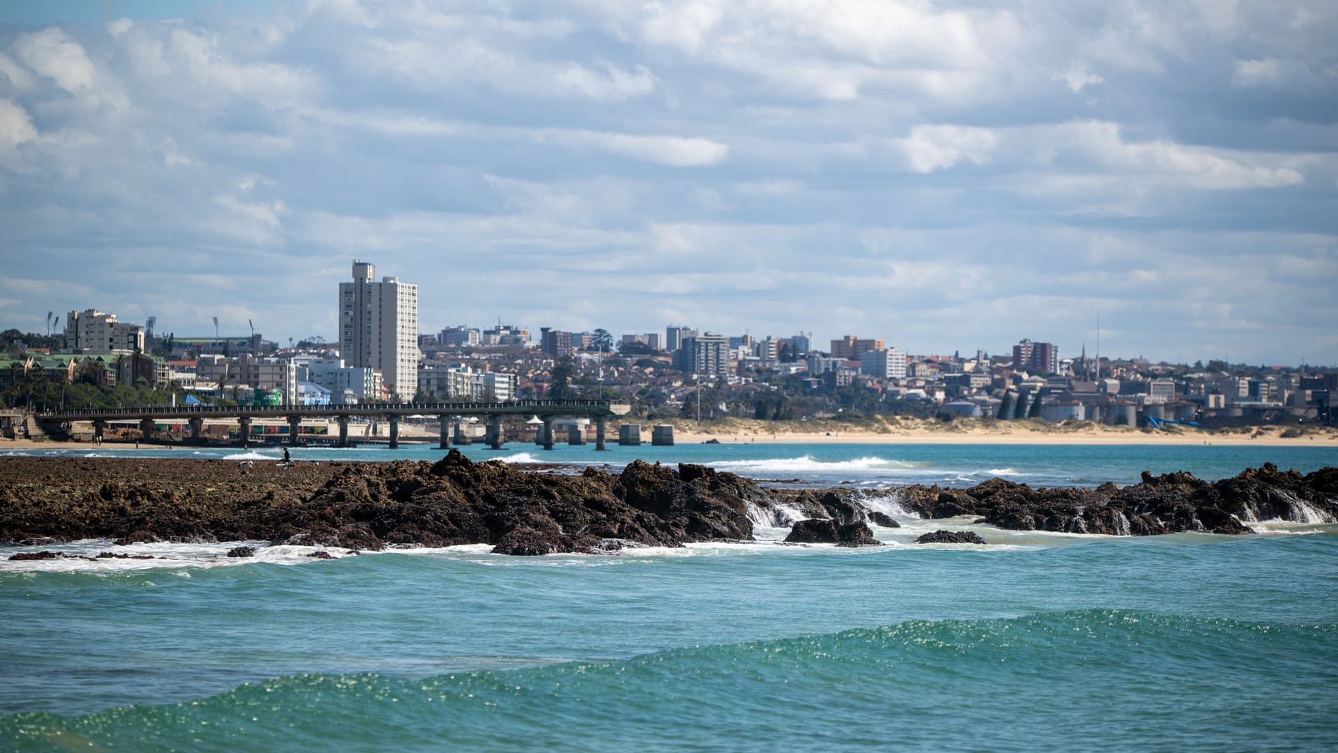 A panoramic shot of the Gqeberha coastline in South Africa, showcasing the city skyline and a pier over the water, with waves crashing on the rocky shoreline in the foreground.