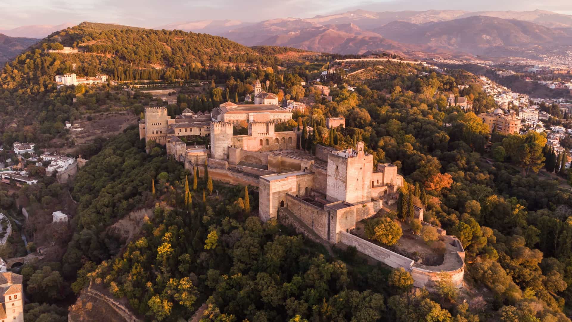 An aerial shot of the magnificent Alhambra palace and fortress complex in Granada, Spain, surrounded by lush green and orange trees with the Sierra Nevada mountains in the background at sunset.