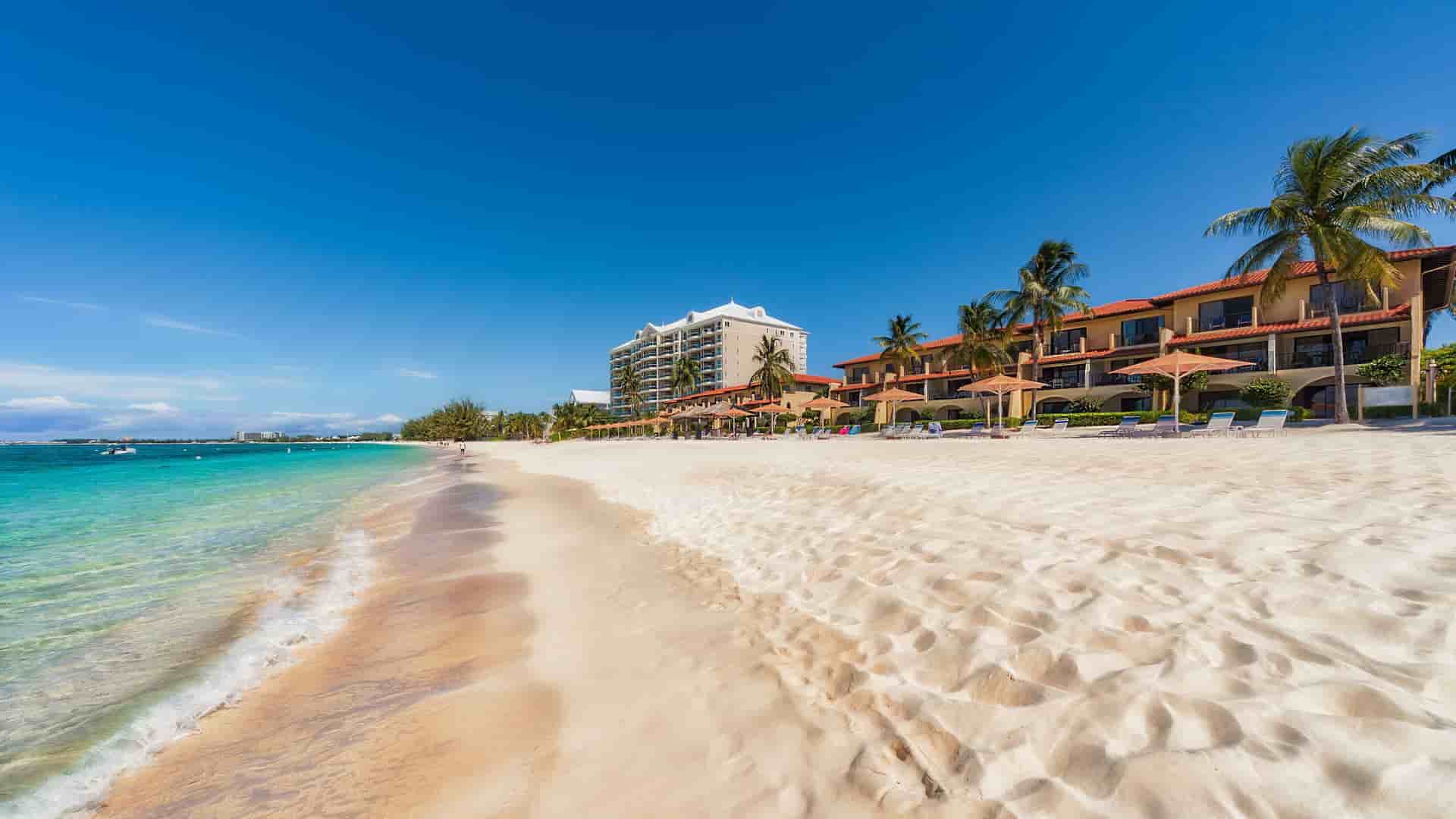 A scenic shot of the stunning Seven Mile Beach in Grand Cayman, with a wide, sandy beach, calm turquoise water, and resorts with palm trees under a beautiful blue sky.