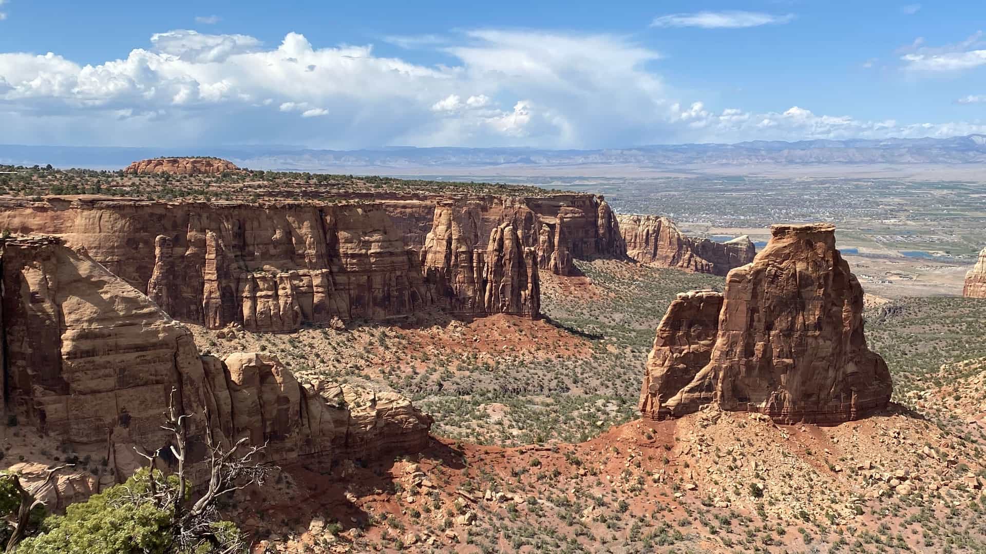 "A scenic panoramic view from the Colorado National Monument near Grand Junction, Colorado, showcasing a desert landscape with dramatic red rock canyons and vast valleys under a bright sky with large clouds. "