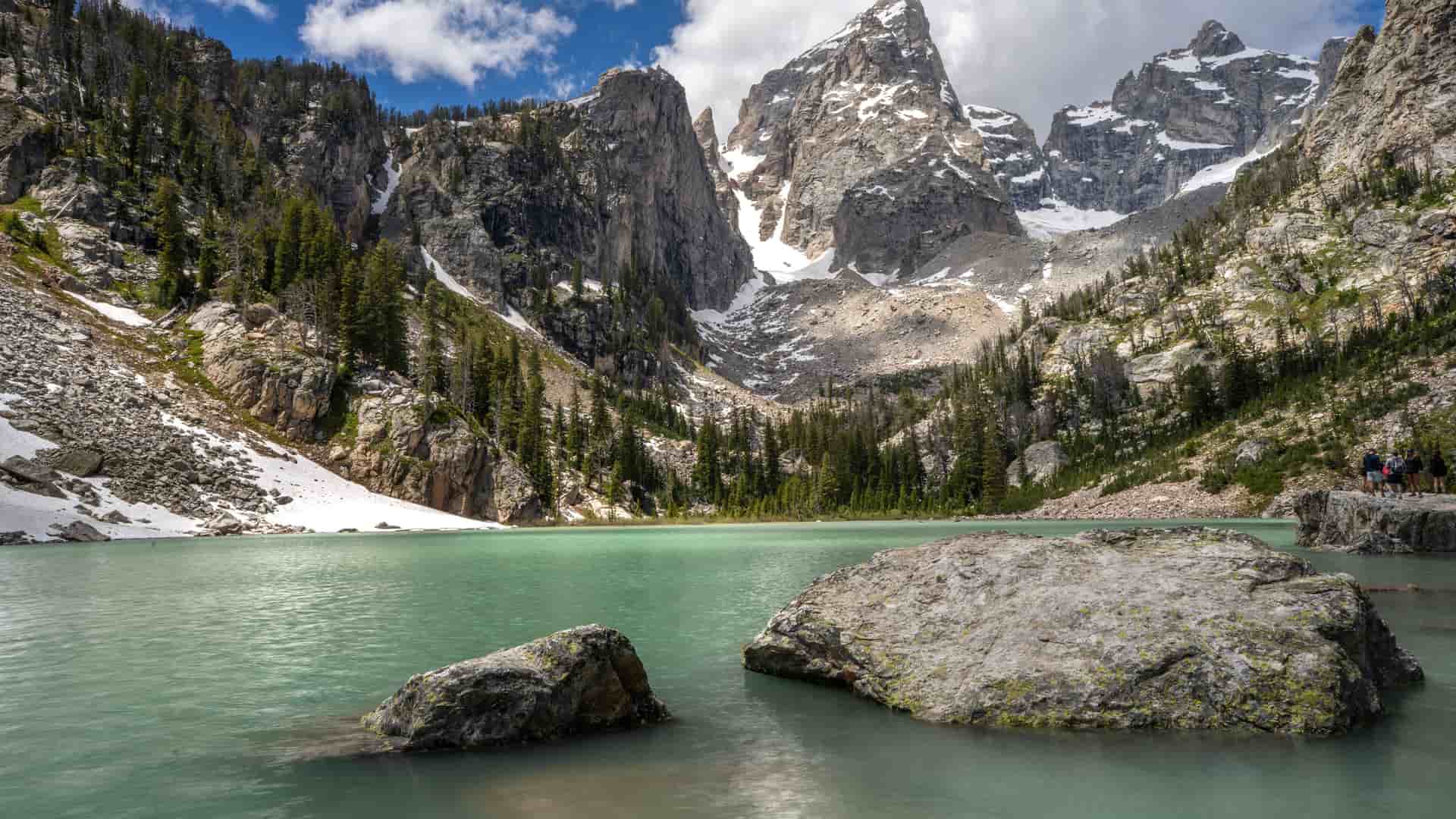 "A beautiful landscape view of Grand Teton National Park, featuring the turquoise waters of Taggart Lake with large boulders in the foreground and the jagged, snow-dusted mountains of the Teton Range in the background. "