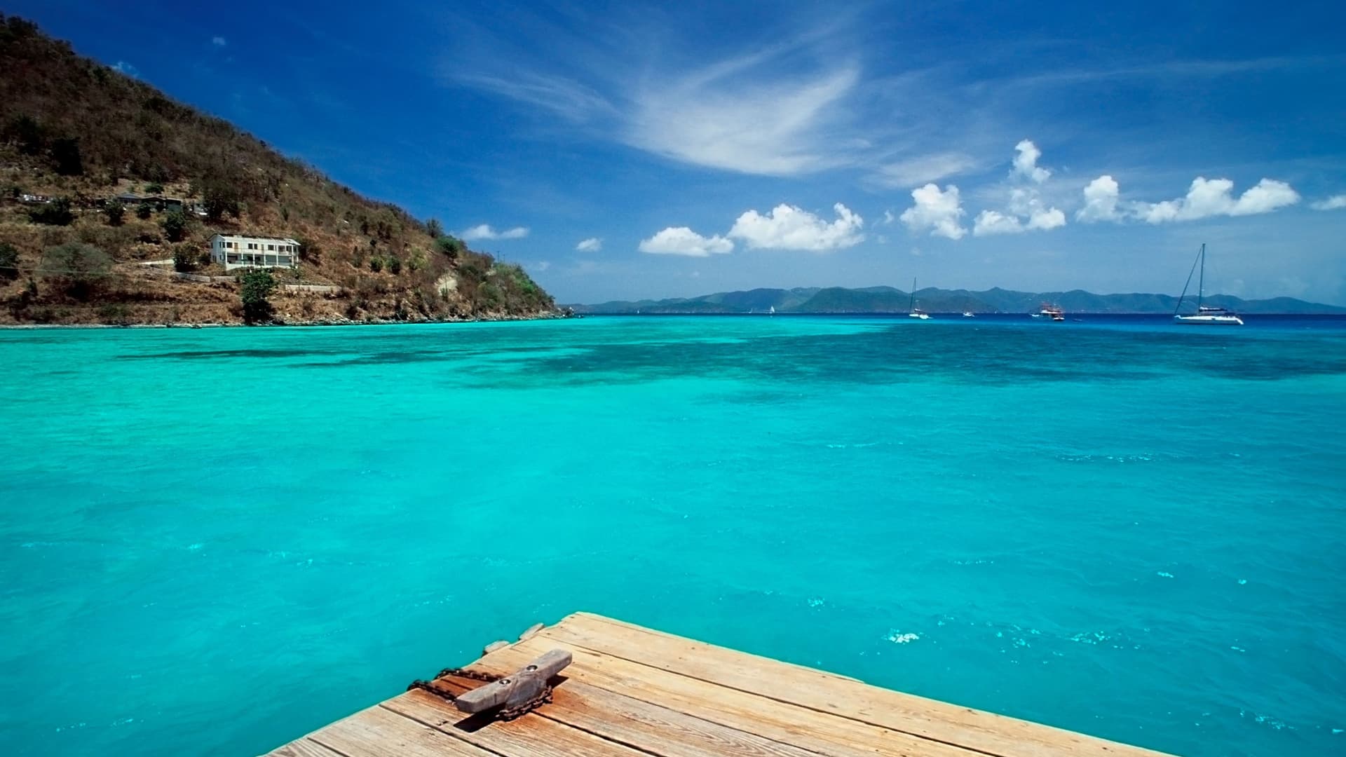 A sunny photo of a wooden dock in Great Harbour, Jost Van Dyke, British Virgin Islands, with sailboats on the turquoise water and a lush green hillside in the background.