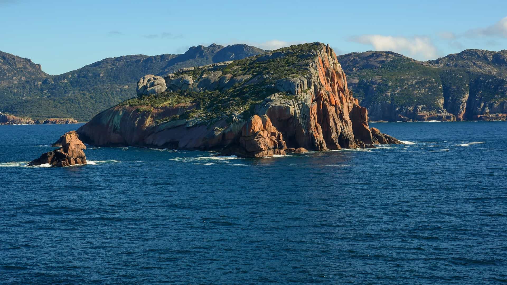 A wide shot of the dramatic, rocky coastline of Freycinet National Park, with a large, distinctively orange-red granite sea stack jutting out of the calm, blue waters of Great Oyster Bay in Tasmania, Australia.