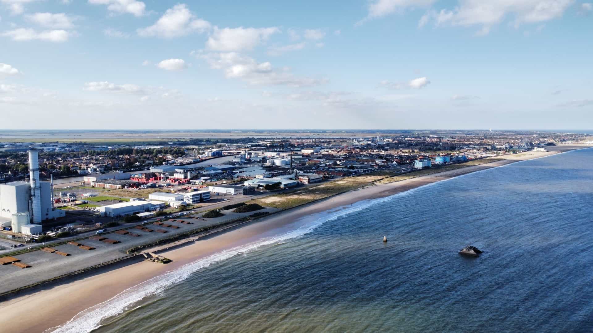 An aerial view of the long, sandy beach and coastline of Great Yarmouth, England, showing the town's industrial and residential areas sprawling inland under a bright blue sky.