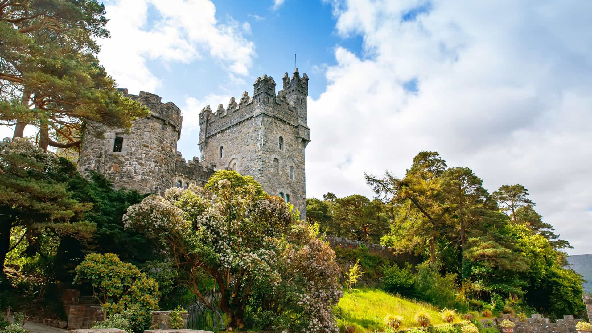 A historic stone castle with a tall tower and crenellated walls, nestled among lush green trees and blooming bushes in Greencastle, Ireland, under a partly cloudy sky.