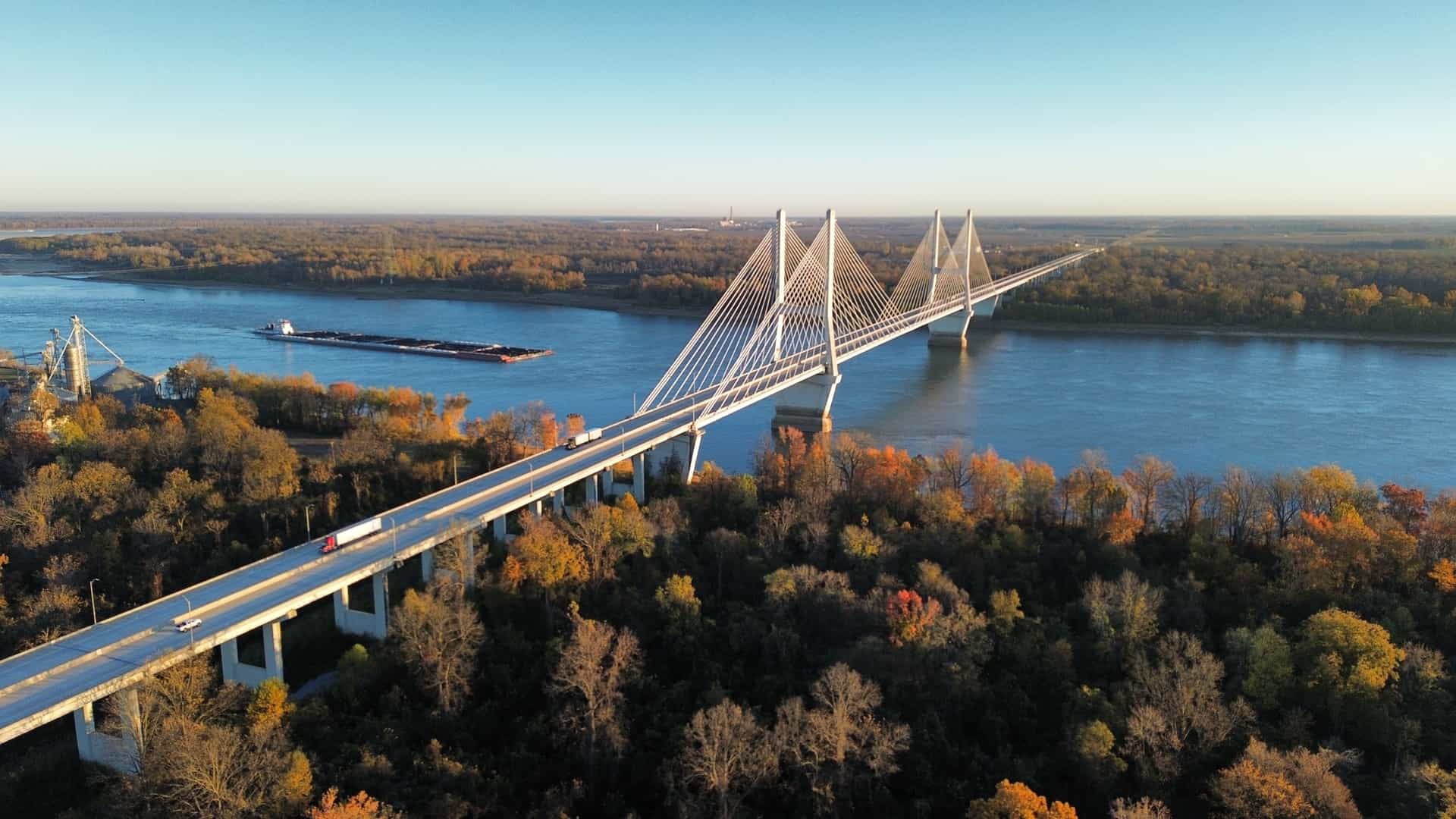 An aerial view of the Greenville Bridge, a modern cable-stayed structure, spanning the wide Mississippi River surrounded by trees with vibrant fall foliage.