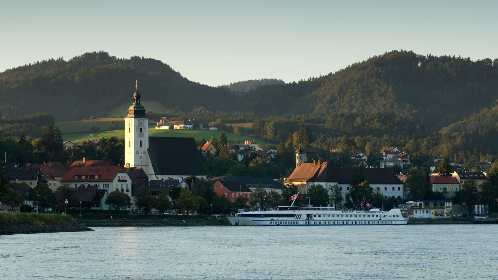 A river cruise ship docked in the picturesque town of Grein, Austria, with a prominent church steeple and residential buildings nestled against lush green hills.