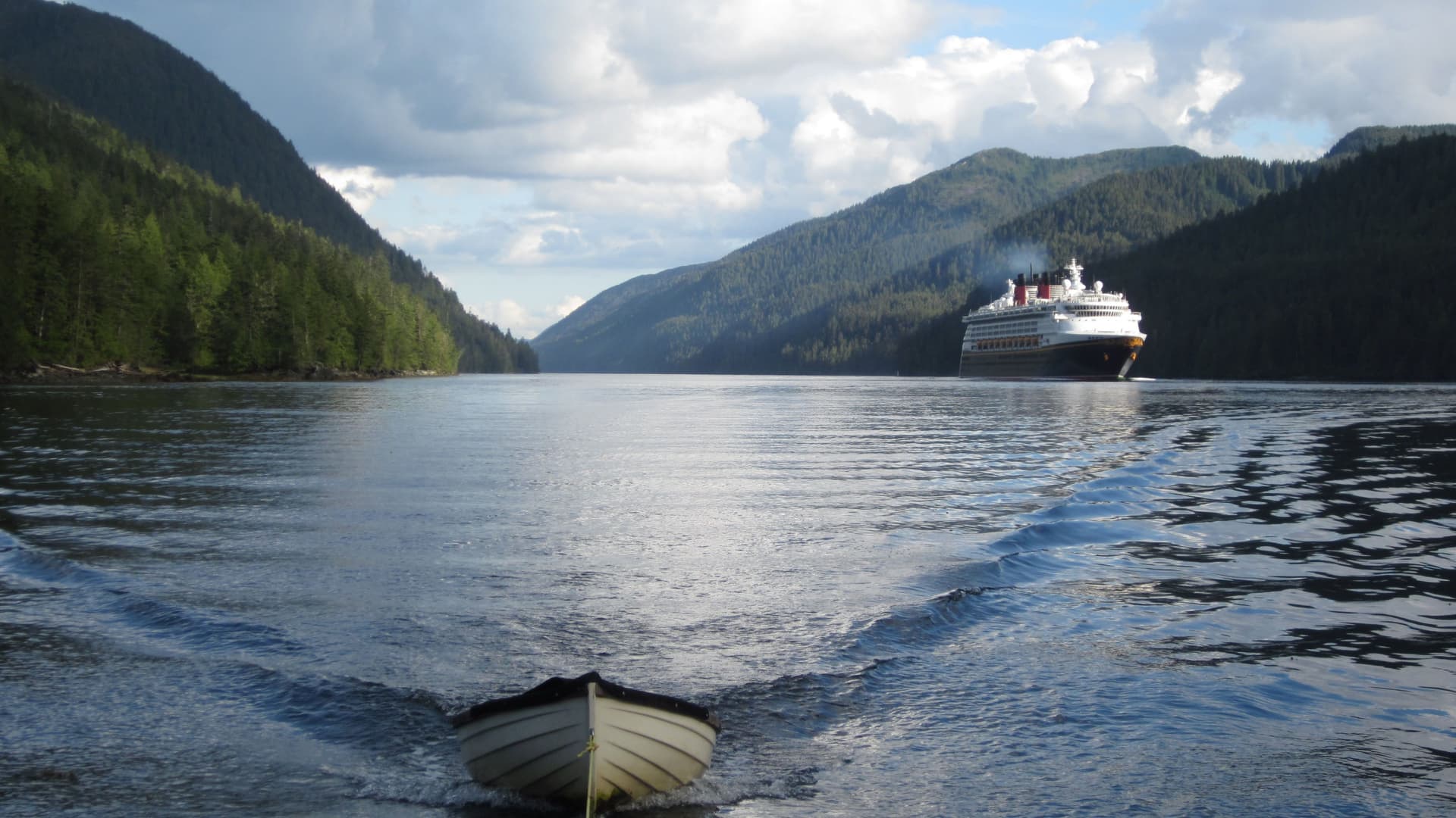 A large cruise ship sailing through the scenic Grenville Channel in British Columbia, with a small boat in the foreground and mountains covered in lush green forests on both sides.
