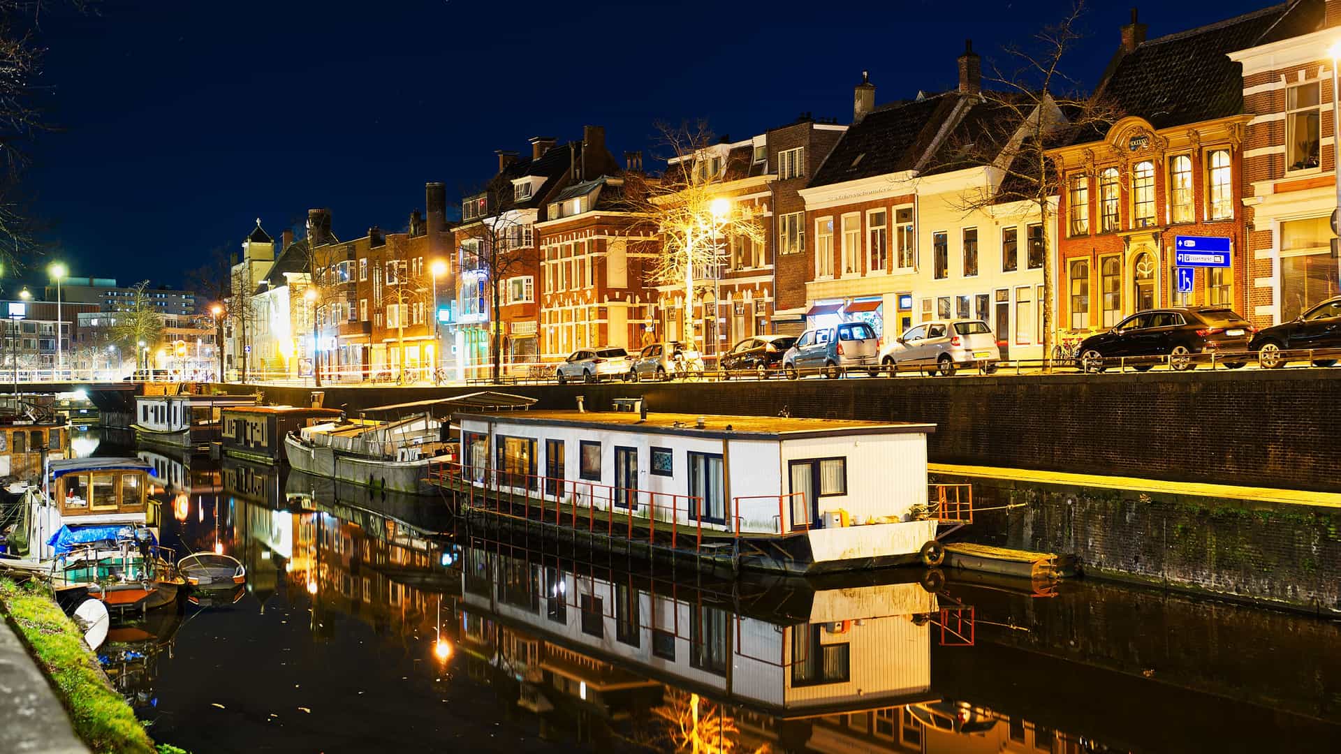 "A vibrant nighttime shot of a canal in Groningen, Netherlands, lined with traditional houses and parked cars, with several illuminated houseboats reflected on the calm water. "