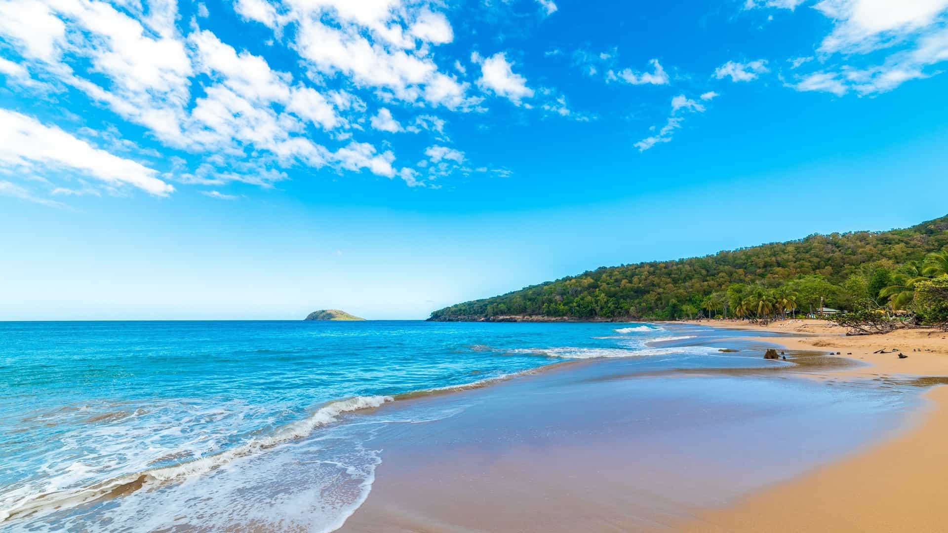 A tropical beach on Guadeloupe with golden sand, bright turquoise water, and a lush, green hillside.