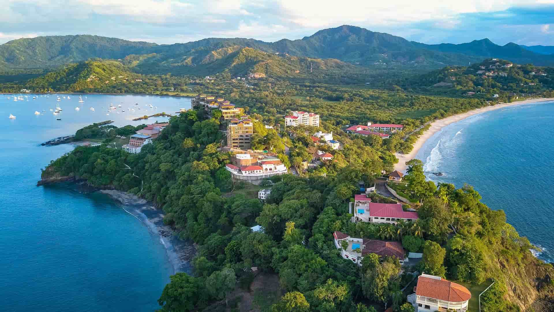 "An aerial view of the coastline of Guanacaste, Costa Rica, showing a verdant peninsula with luxury resorts and homes, with a white sand beach and boats moored in the clear blue water. "