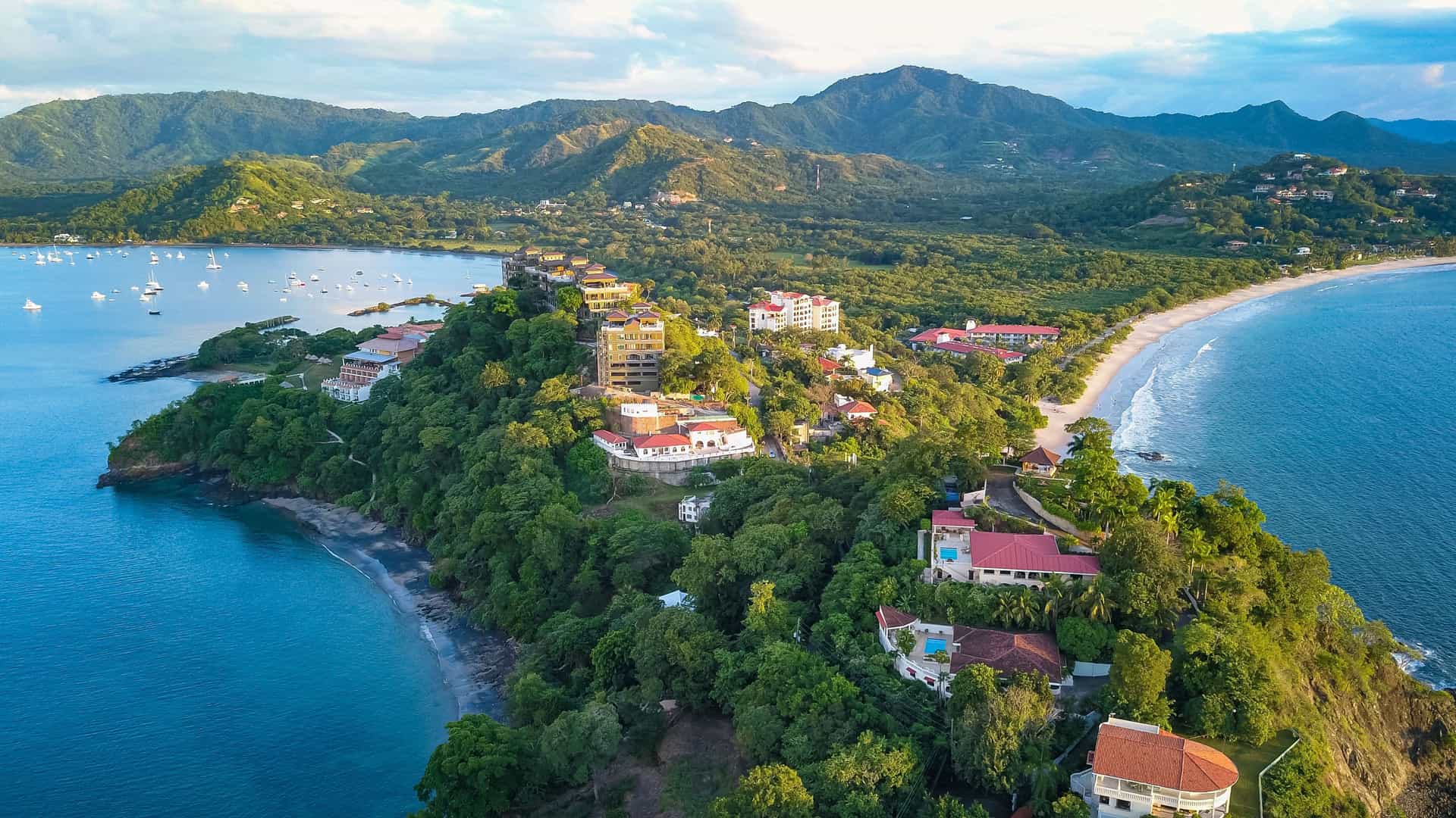 "An aerial view of the coastline of Guanacaste, Costa Rica, showing a verdant peninsula with luxury resorts and homes, with a white sand beach and boats moored in the clear blue water. "