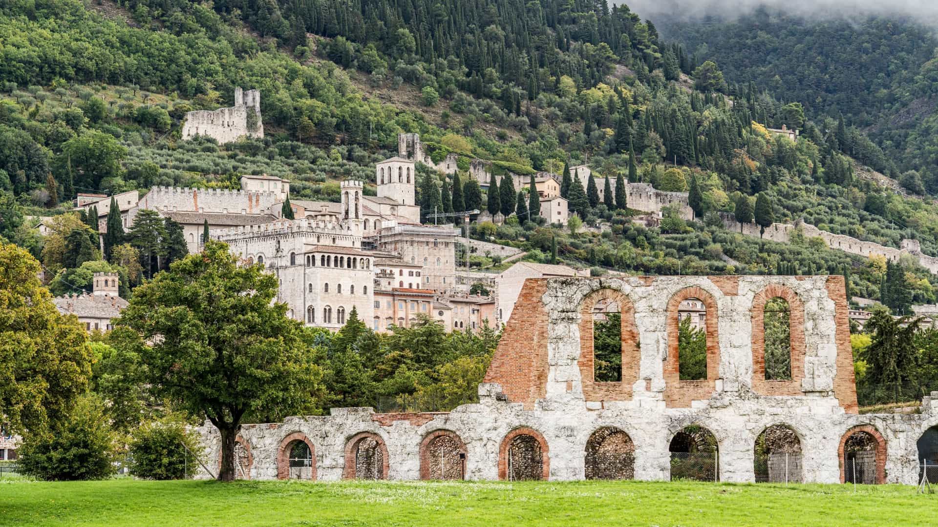 Ruins of a Roman archway and red brick structure in the foreground, with the medieval hill town of Gubbio, Umbria, Italy, visible on the slope of Mount Ingino behind a grassy field.