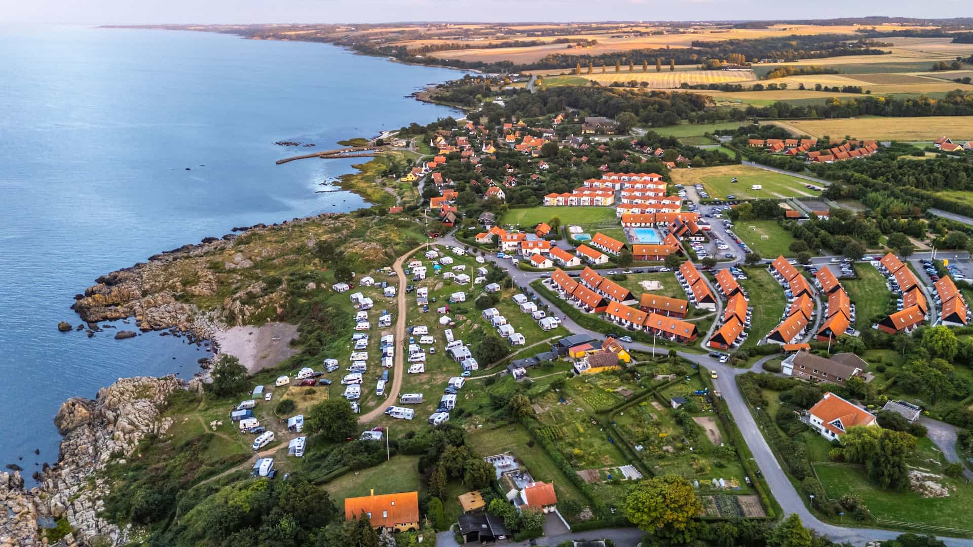 An aerial view of the scenic coastline of Gudhjem, Bornholm Island, Denmark, showing a caravan campsite and the town's red-roofed buildings nestled along the rocky shore.